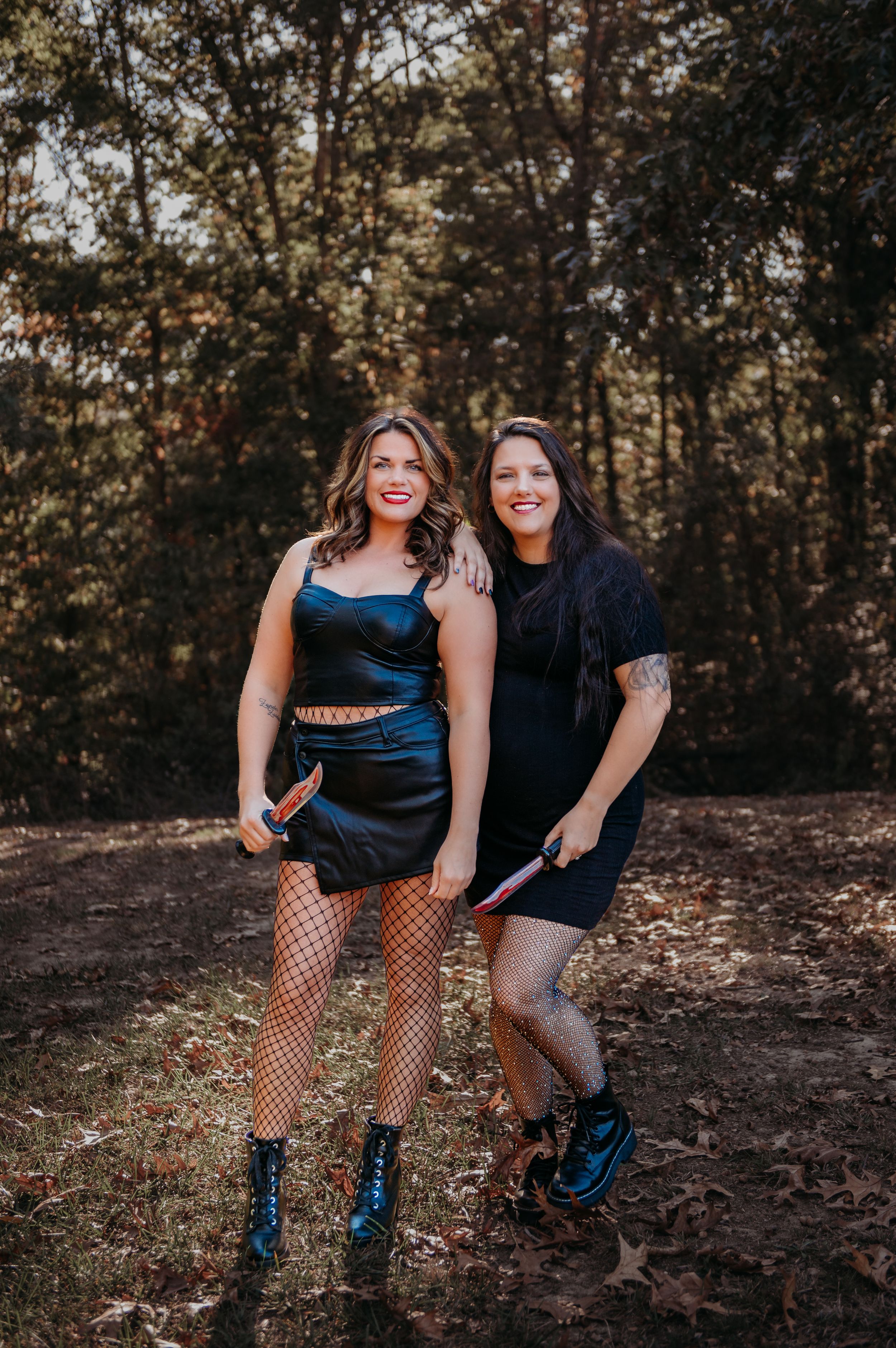 Two people in dark outfits and fishnet stockings pose in wooded setting during sunset.
