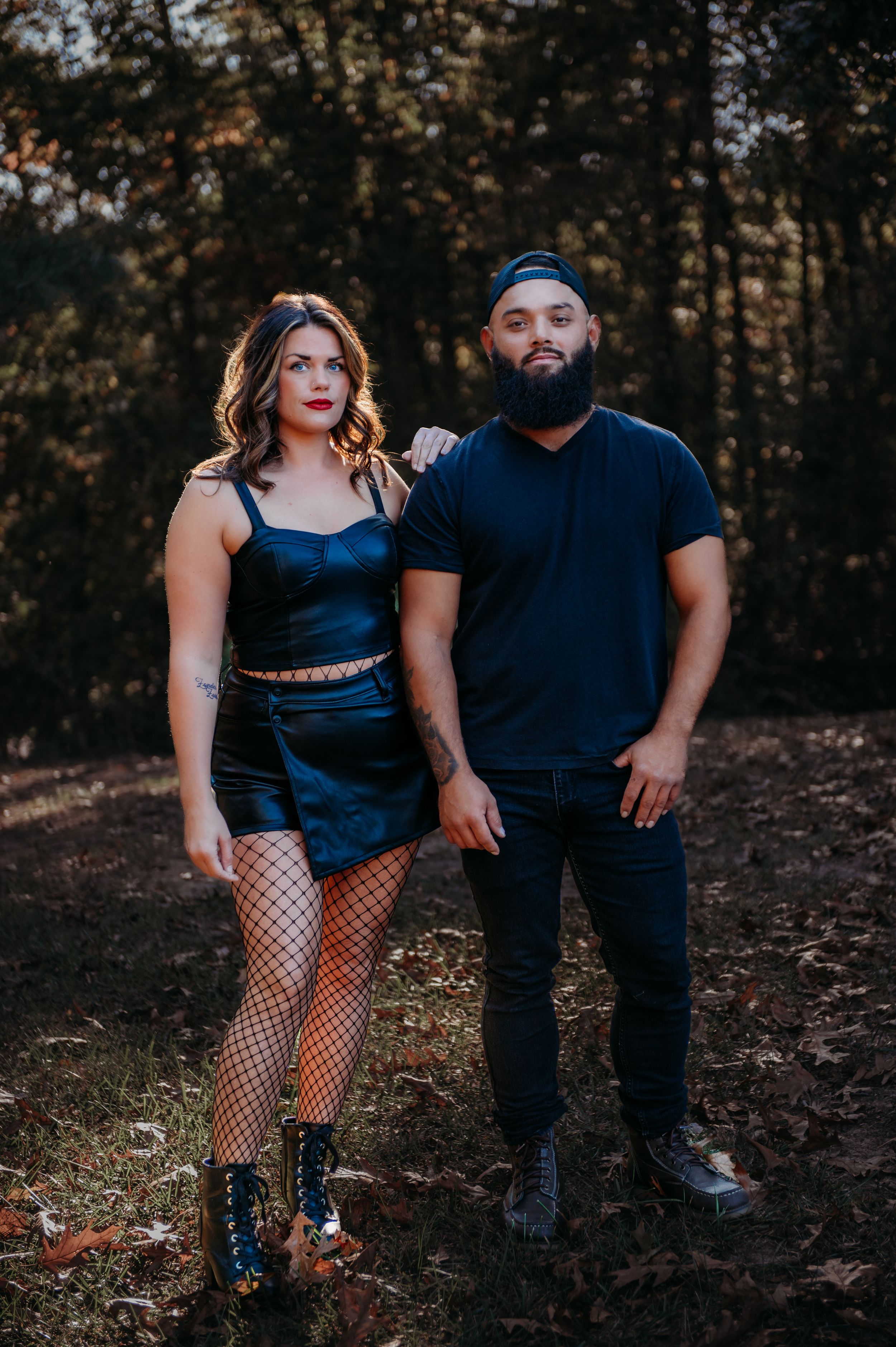 Couple in dark clothing poses together in autumn forest setting.