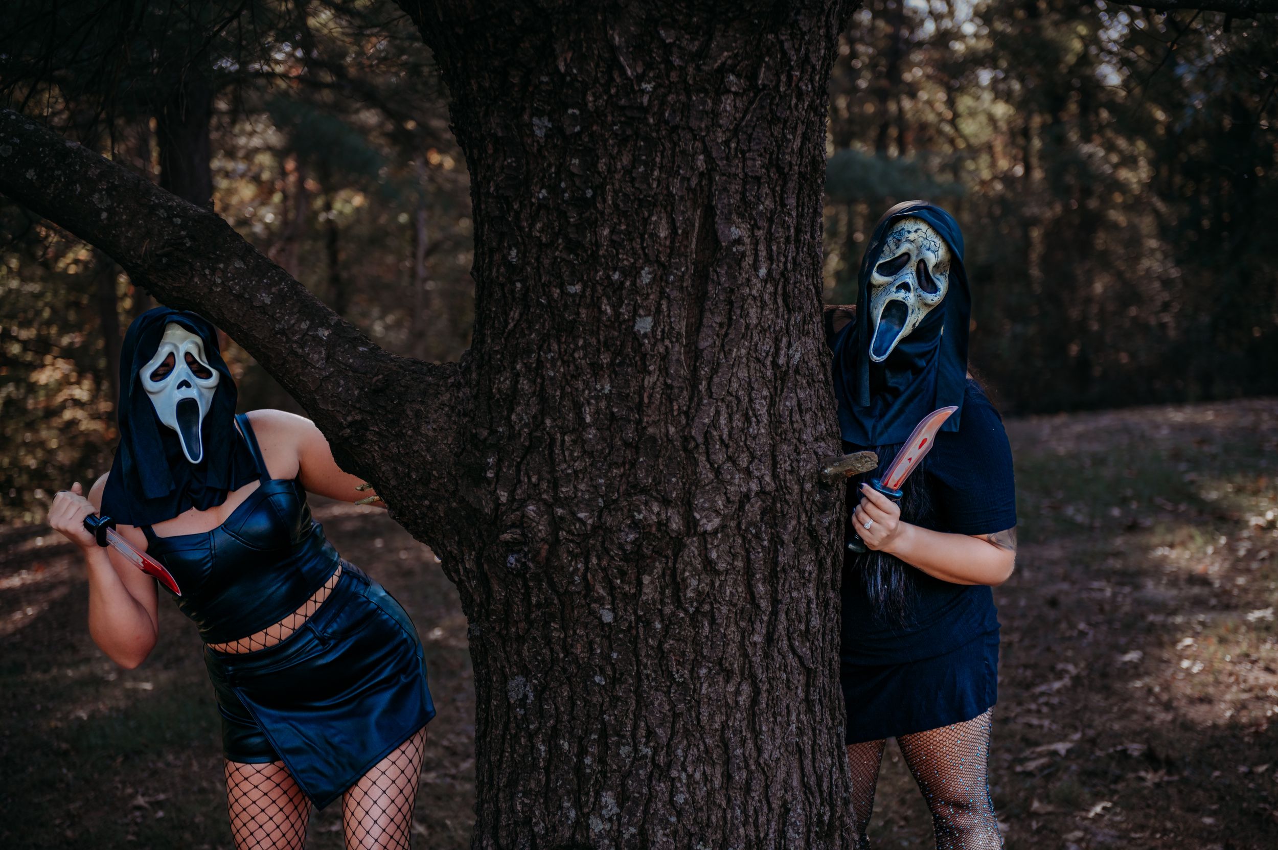 Two figures in blue dresses and ghost masks pose playfully behind a tree in an autumn forest.