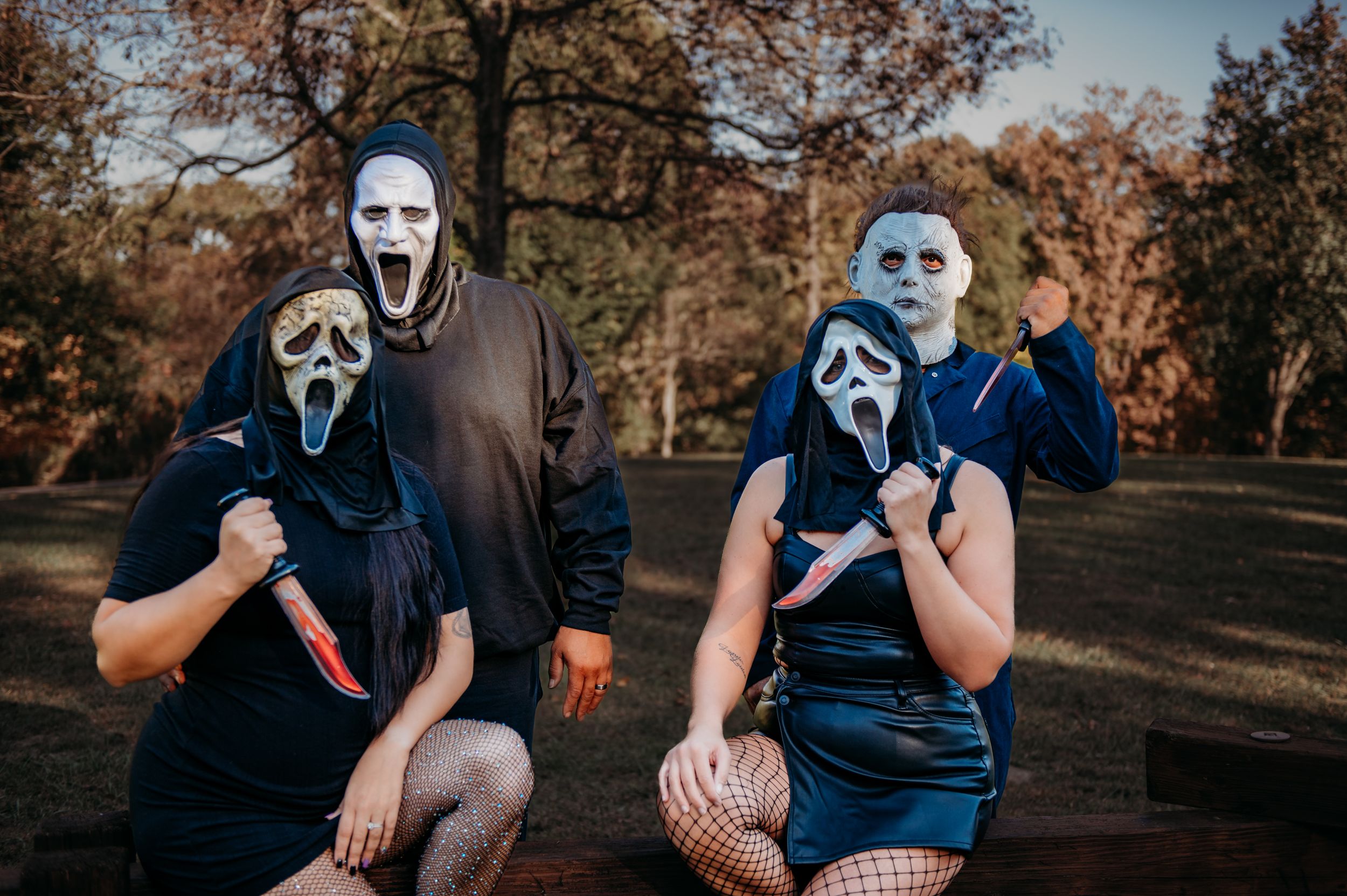 A group poses outdoors wearing horror movie masks and casual clothing in an autumn setting.
