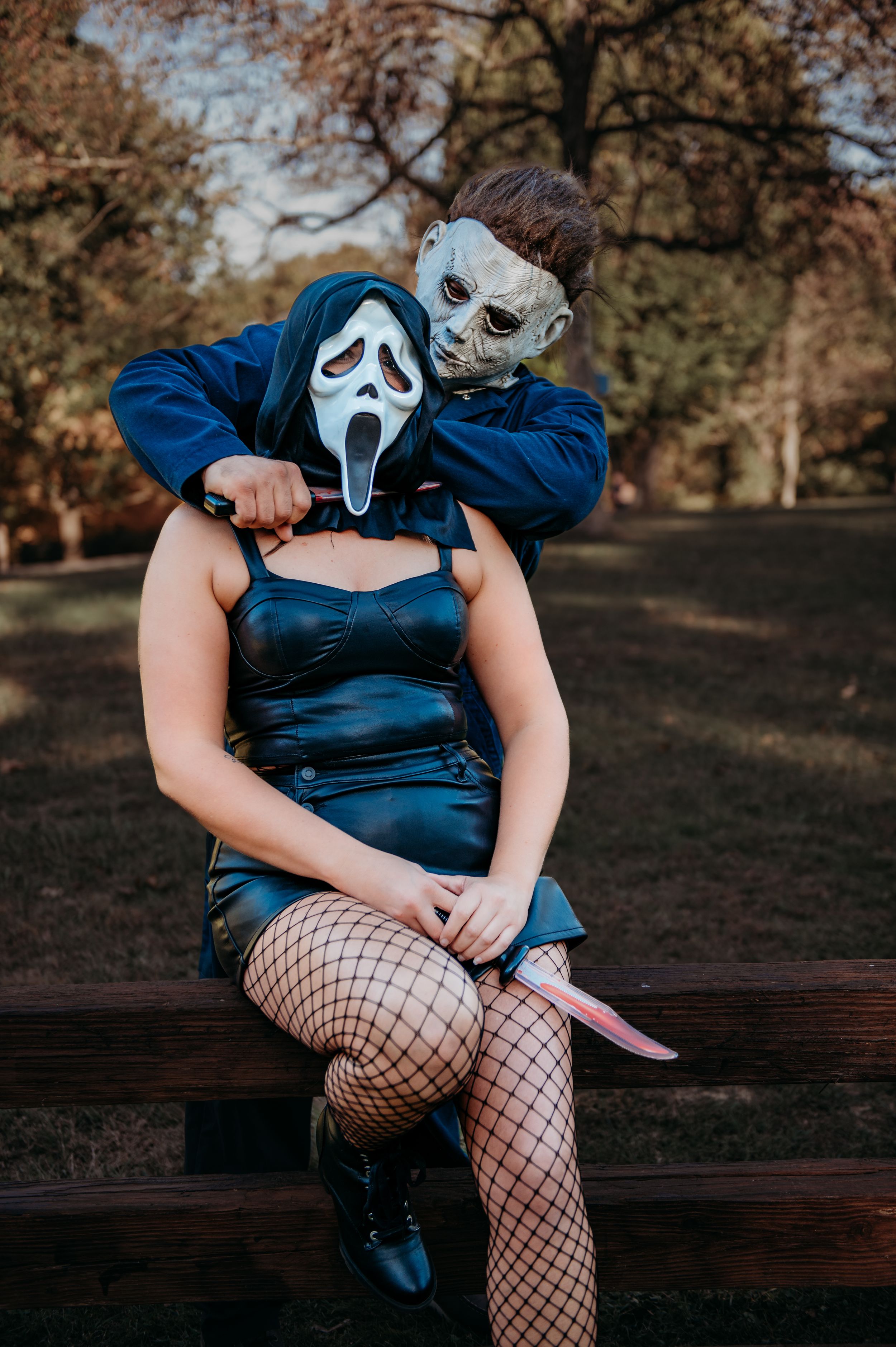 Two people in horror movie masks pose together on a wooden bench in a park setting.