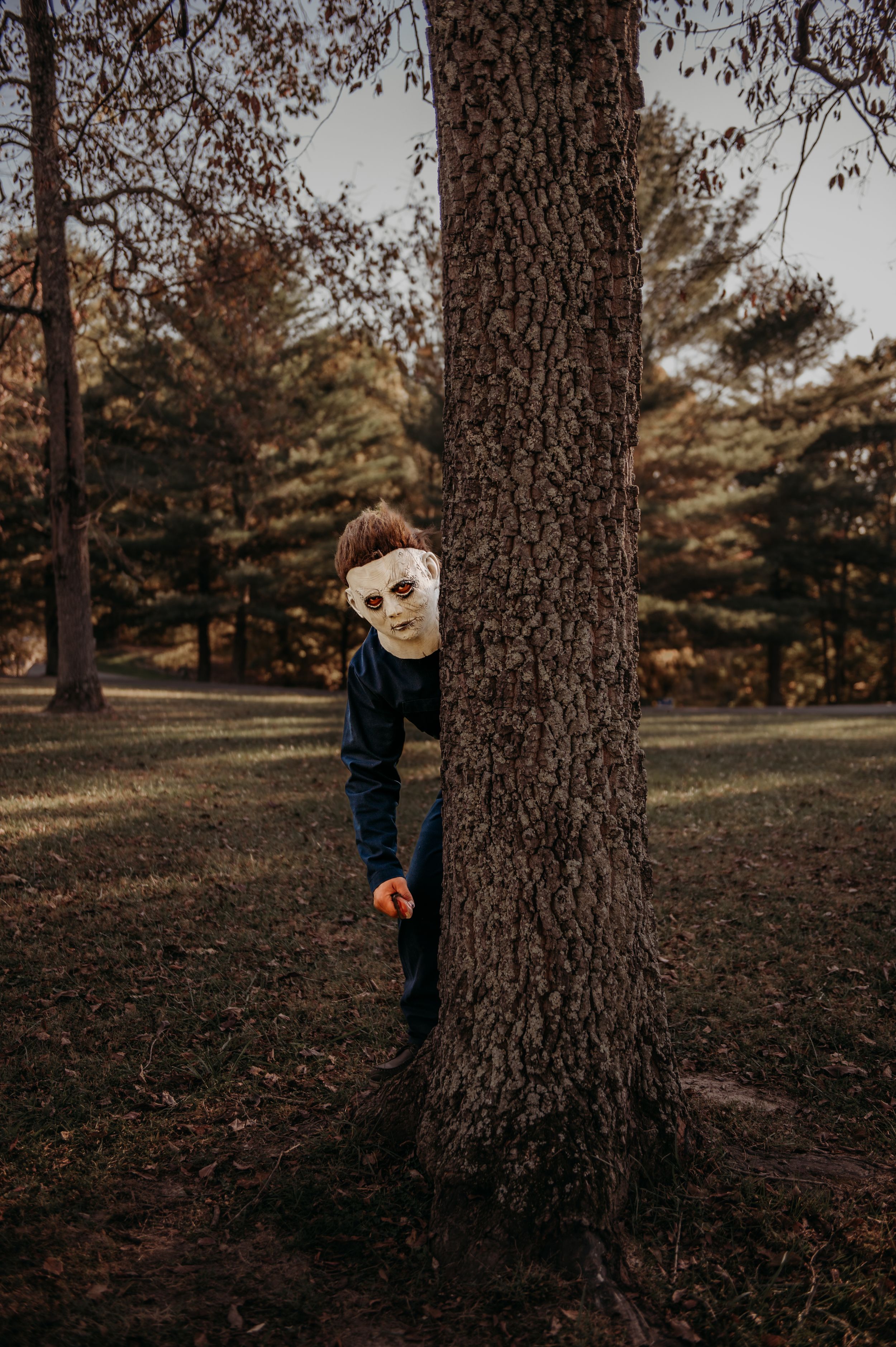 A figure in a classic horror movie mask peers mysteriously from behind a large tree trunk.