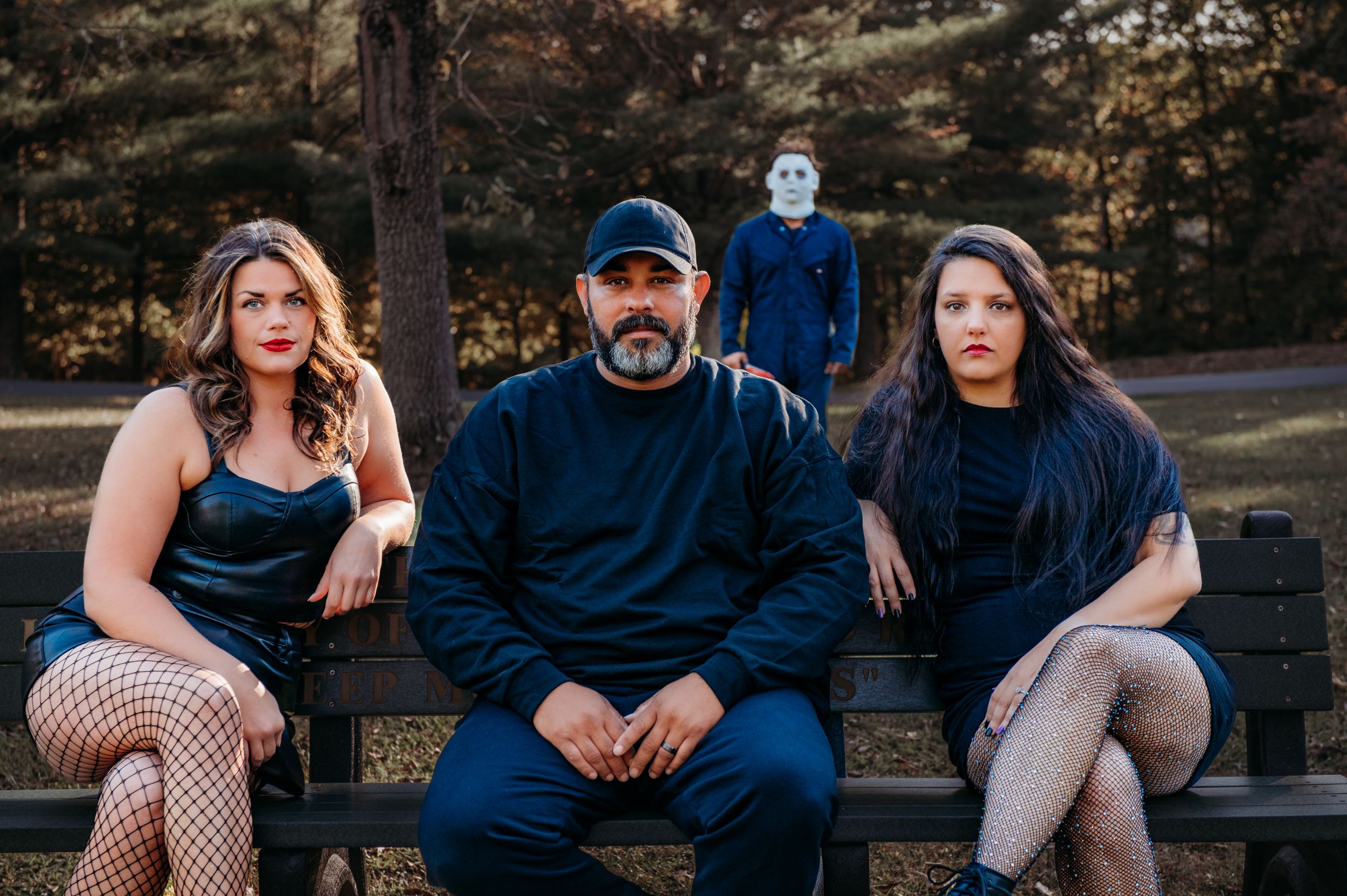 Three people sit on a park bench with a mysterious figure in a mask lurking behind them.