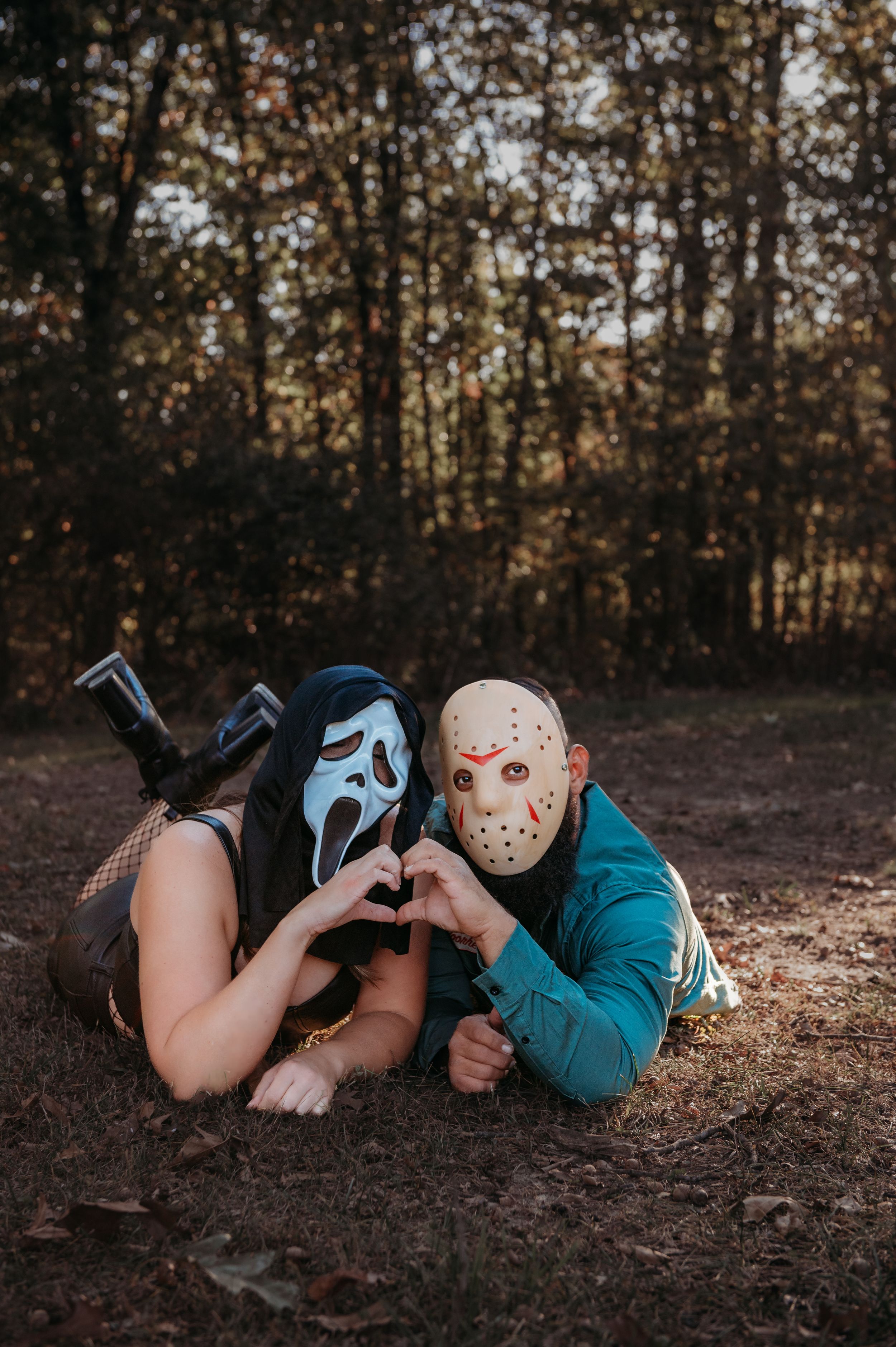 Two people wearing horror movie masks make a heart shape while lying on autumn ground.