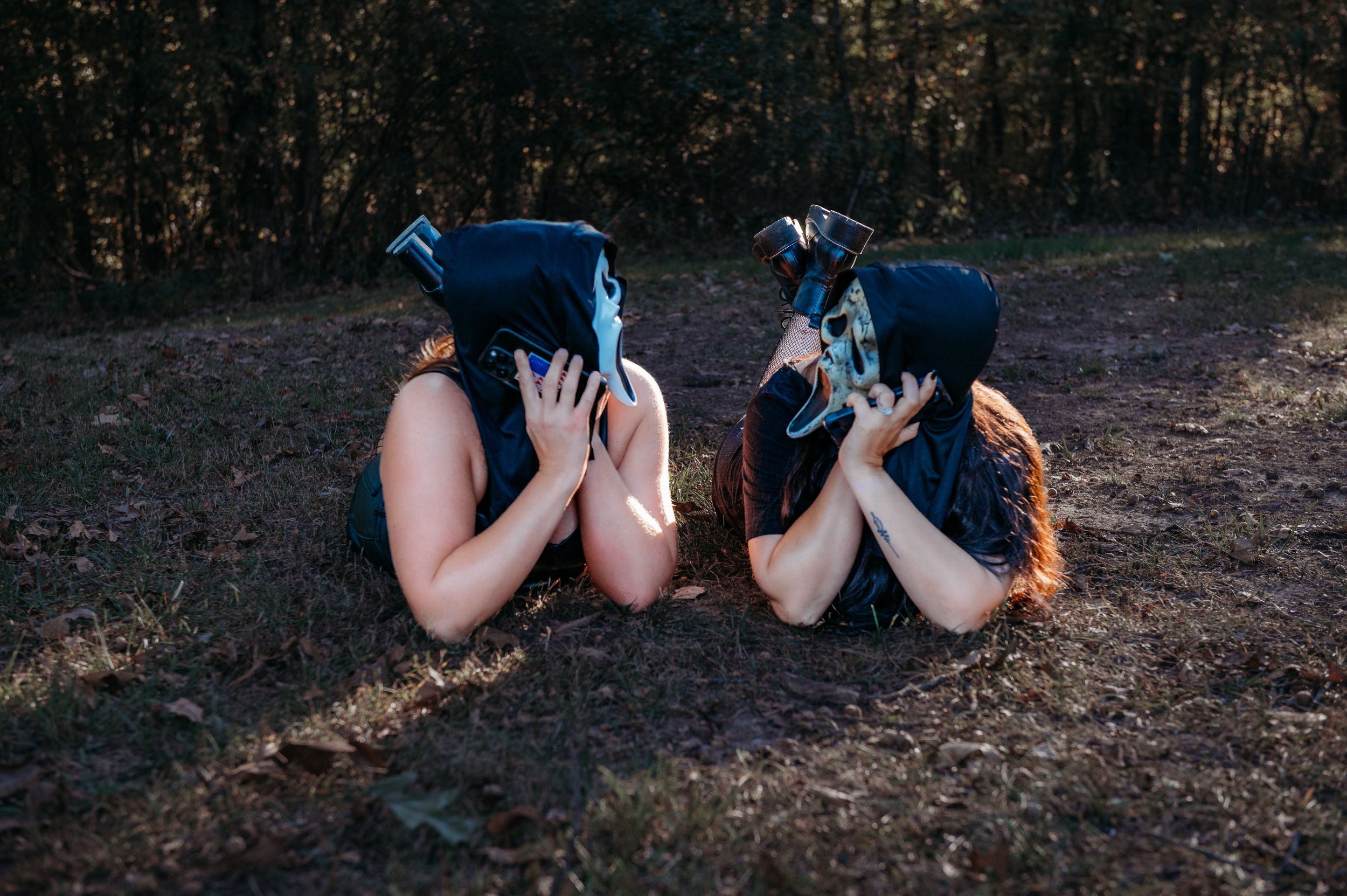 Two people in blue dresses lie on autumn ground covering their faces with ghostface masks.