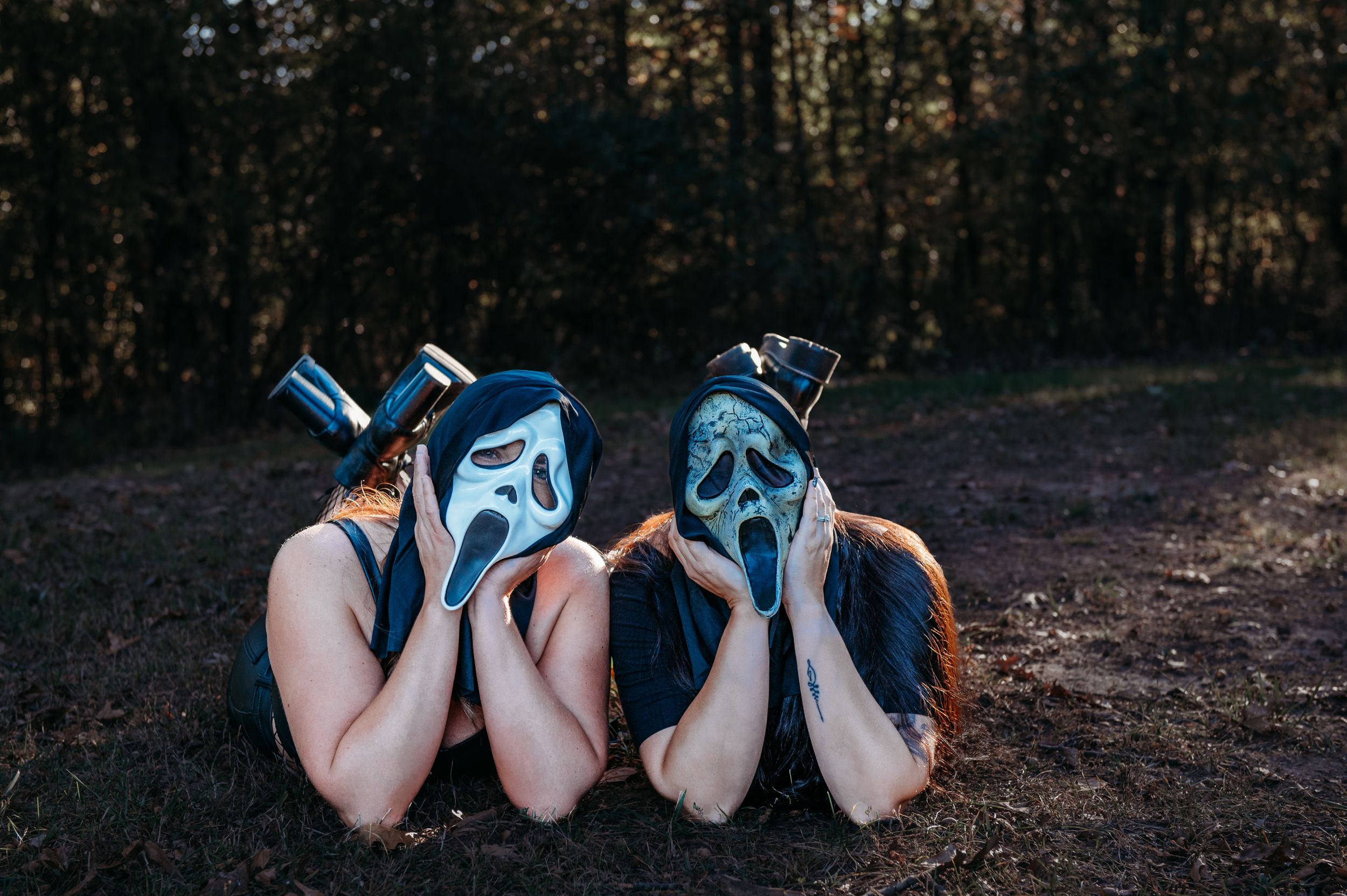 Two people wearing ghostface masks lie on fallen leaves while taking photos.