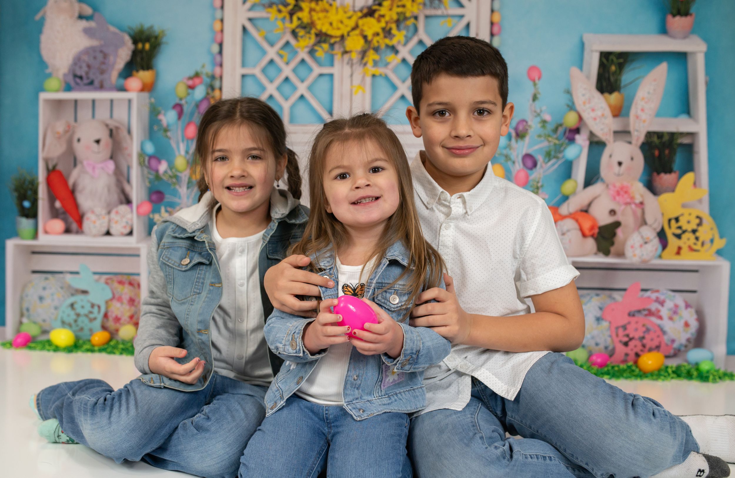 Three children sitting together with Easter decorations and pink bunny plushies in the background.