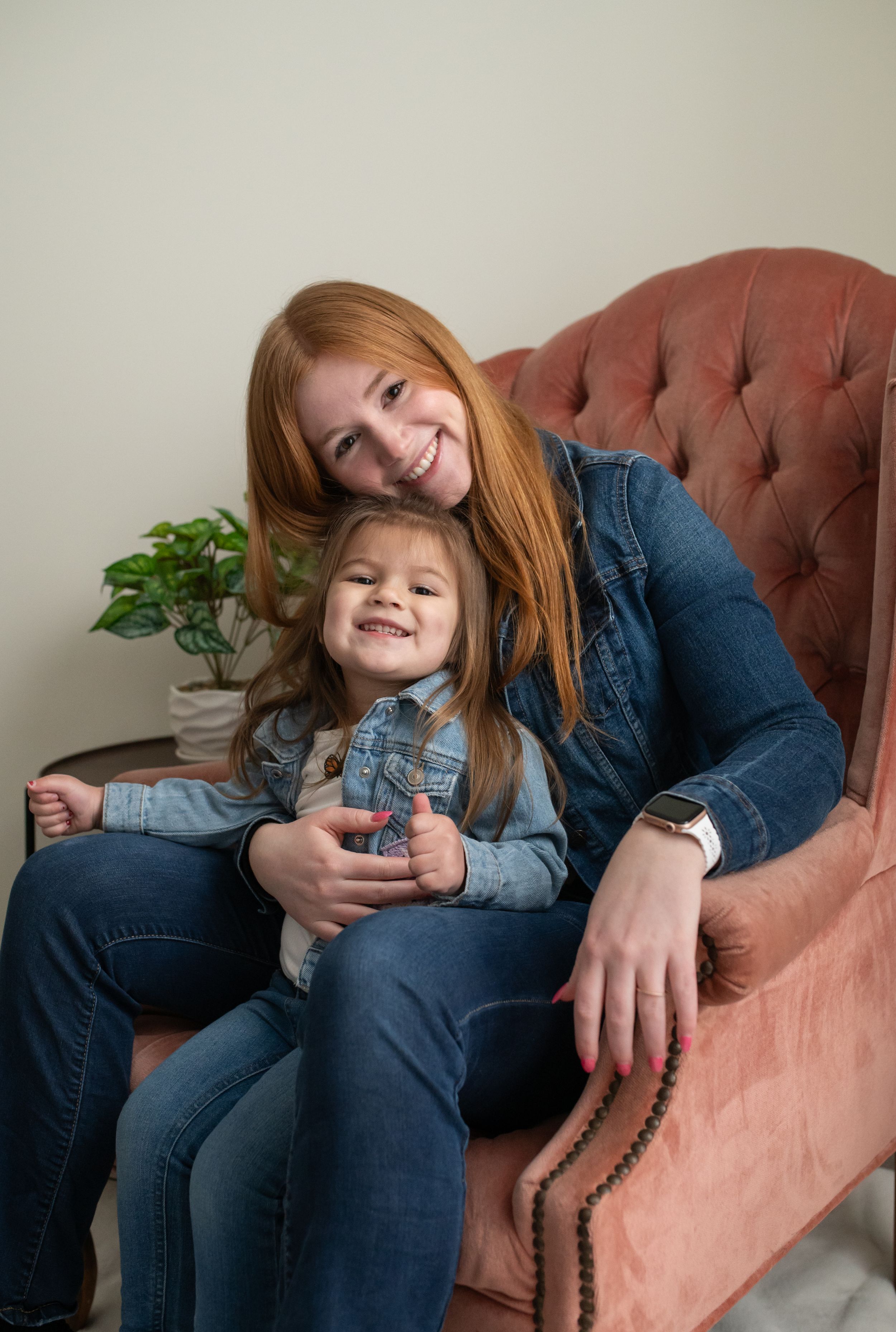 Mother and daughter in matching denim outfits sitting together on a pink velvet armchair.