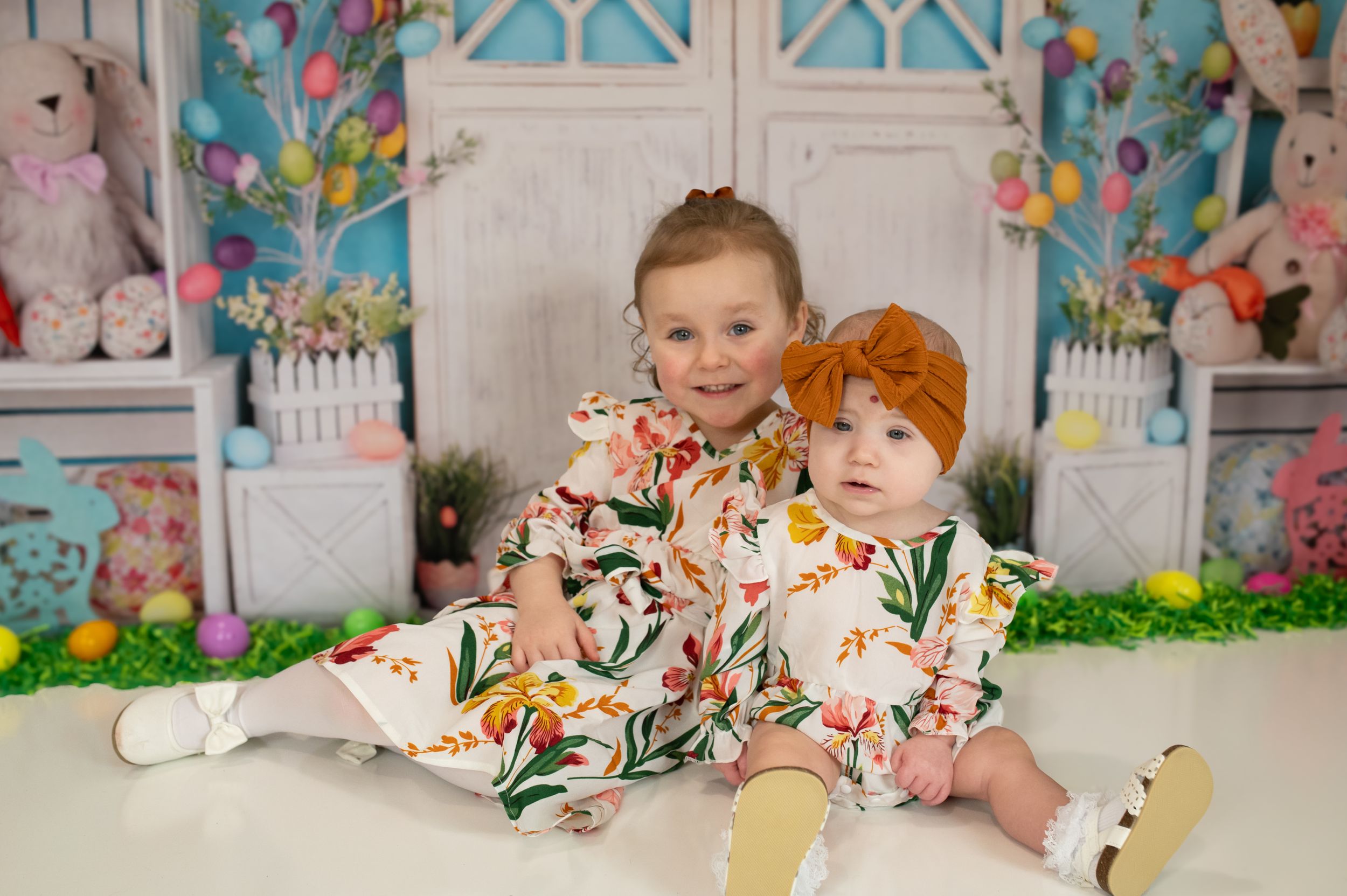 Two siblings in matching floral outfits pose against an Easter-themed backdrop with bunnies.