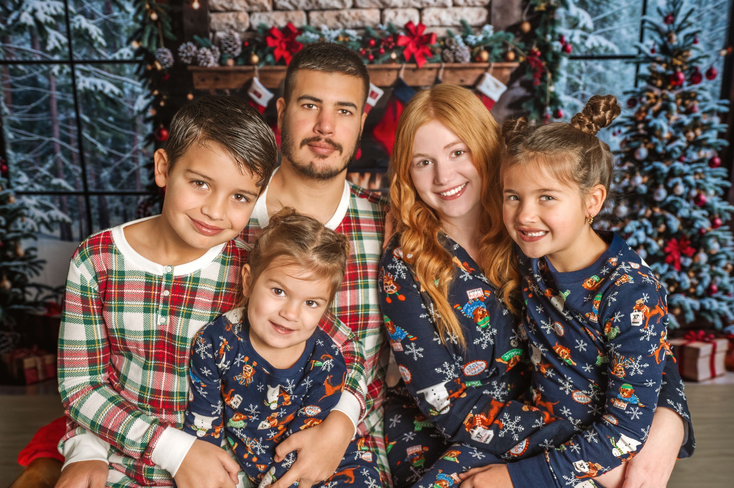 Family wearing matching Christmas pajamas pose in front of decorated fireplace.