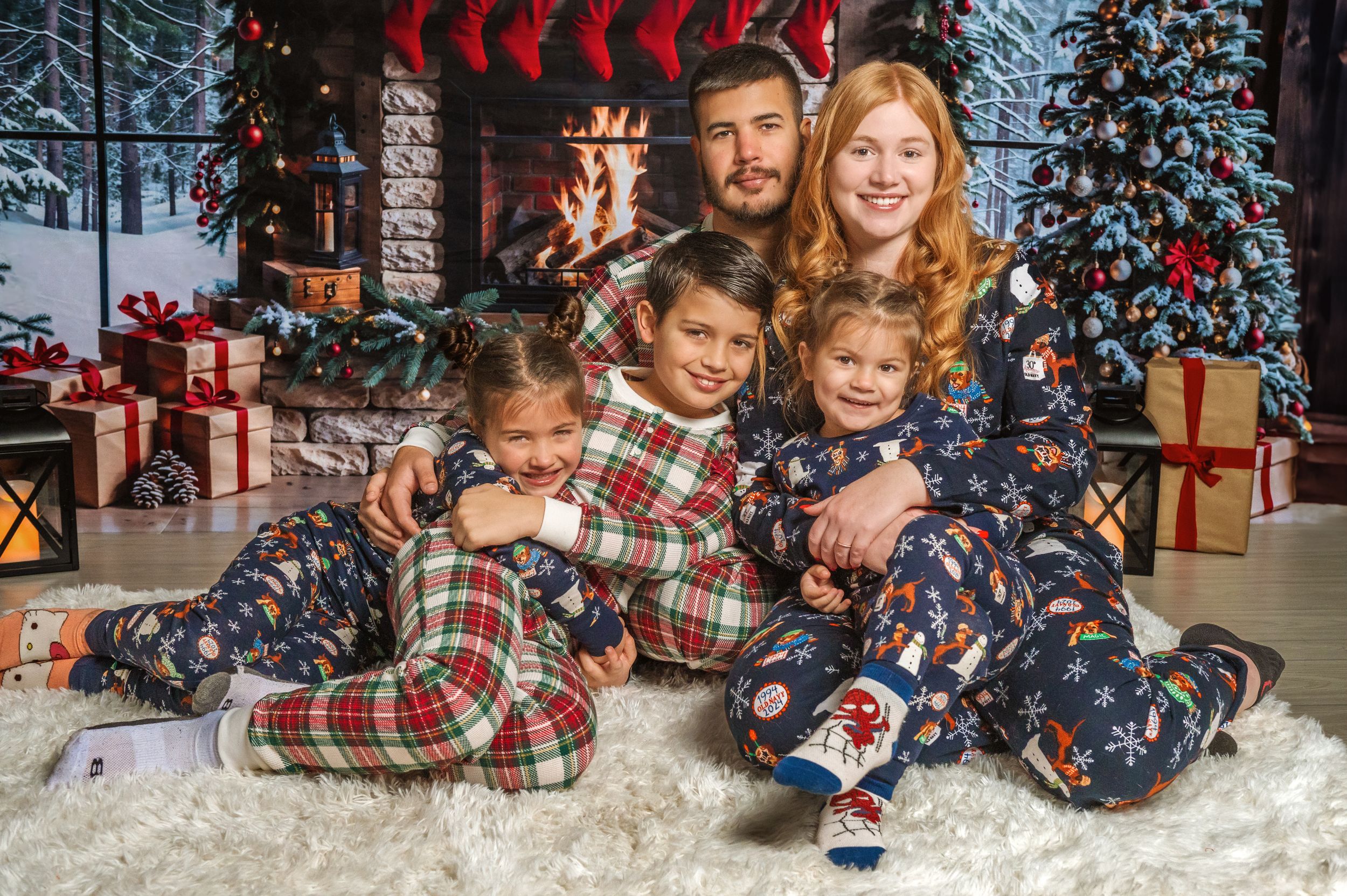 Family in coordinating Christmas pajamas cuddle on white rug by decorated fireplace.