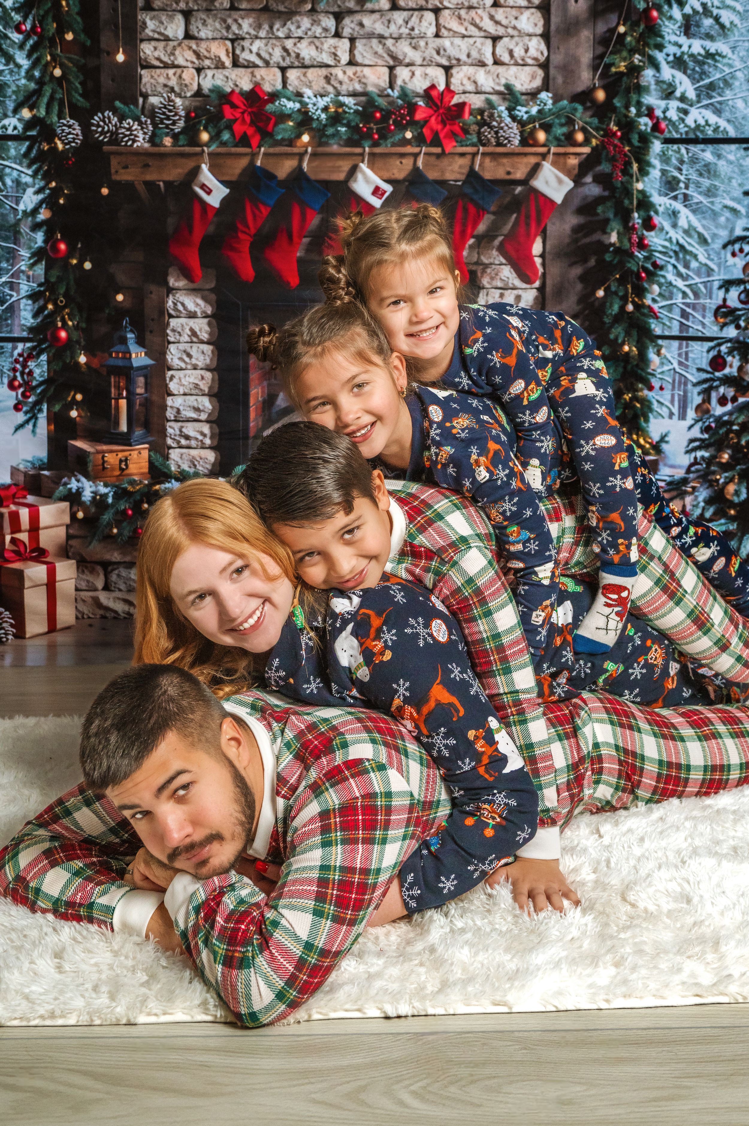 Family in matching holiday pajamas stacked playfully by Christmas fireplace.
