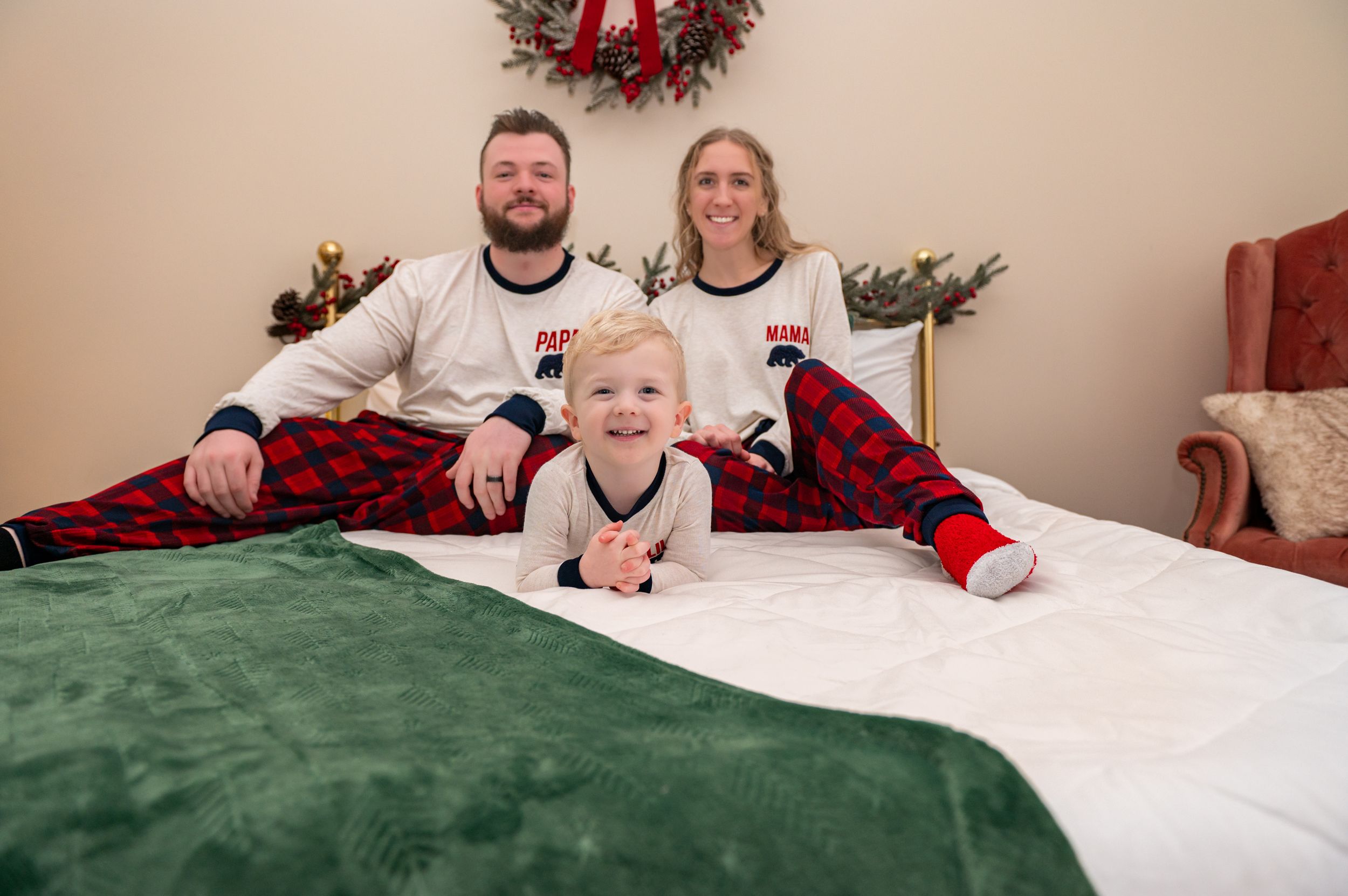 A family wearing matching holiday pajamas poses on a white bed with festive decorations and wreaths above.