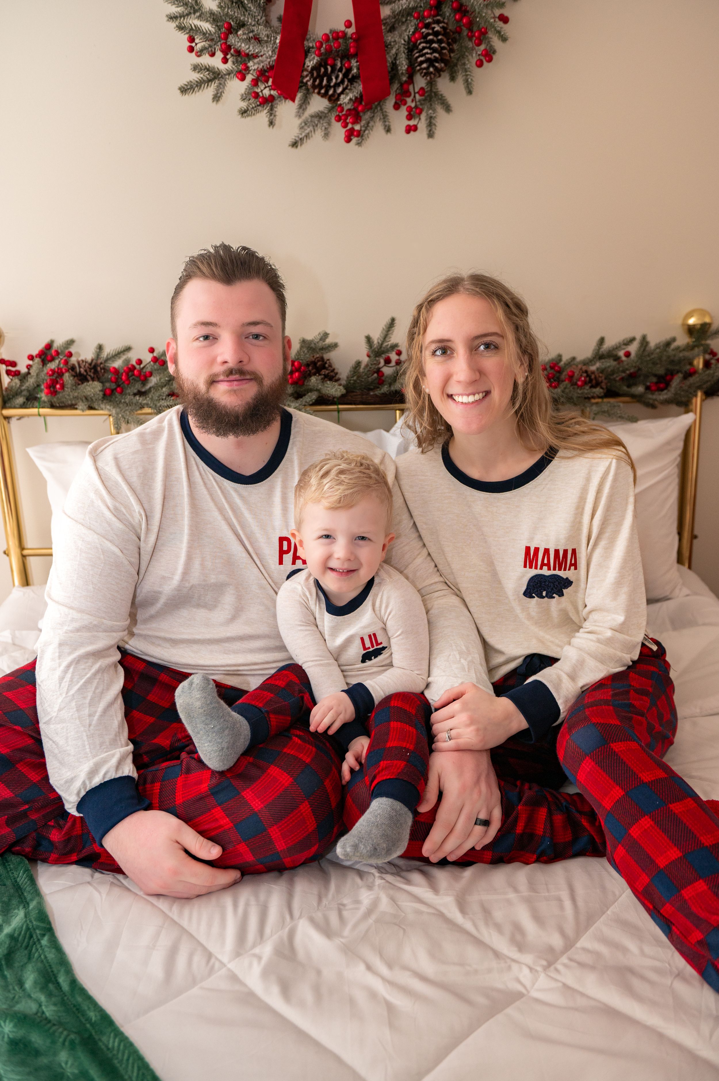 A family in coordinated white and plaid Christmas pajamas relaxes on a bed with holiday decor.