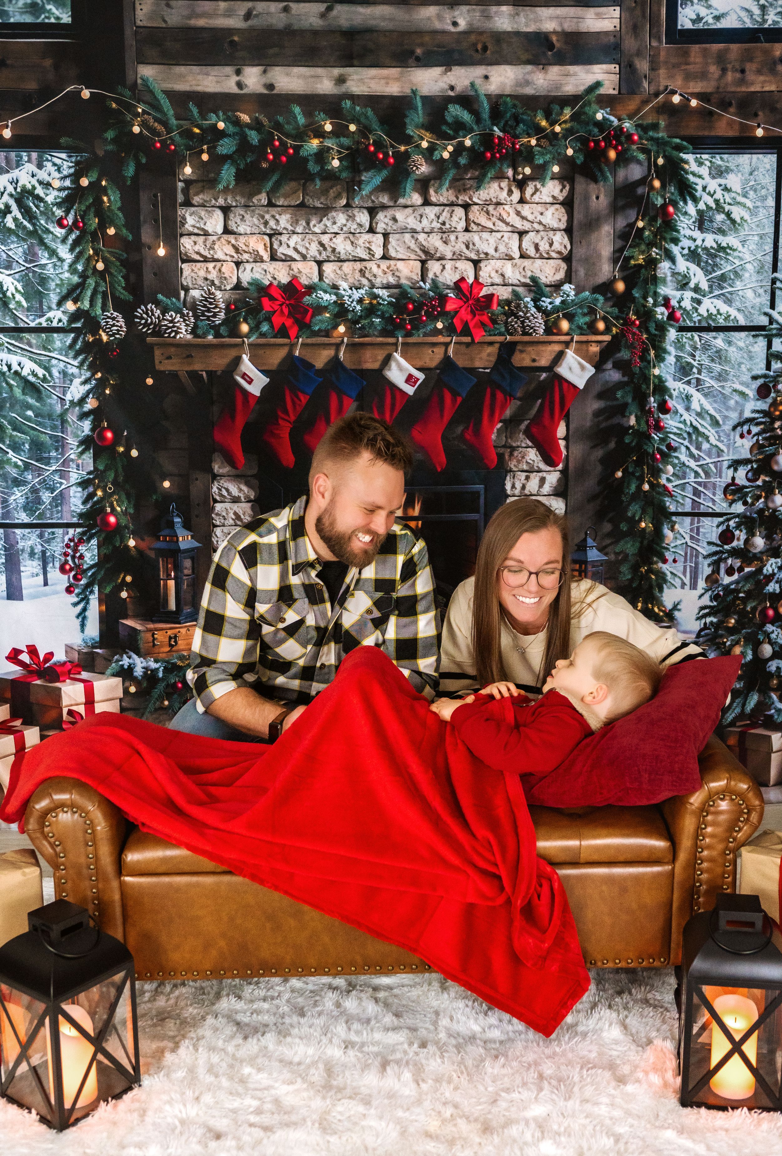 Family lying on brown leather couch with red blanket surrounded by holiday decorations and twinkling lights.