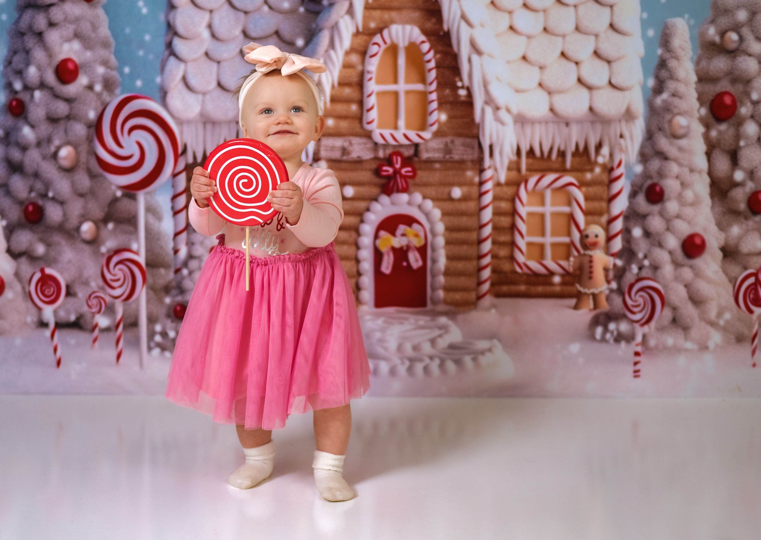 Small child in pink dress poses in front of whimsical candy-themed holiday backdrop with gingerbread house.