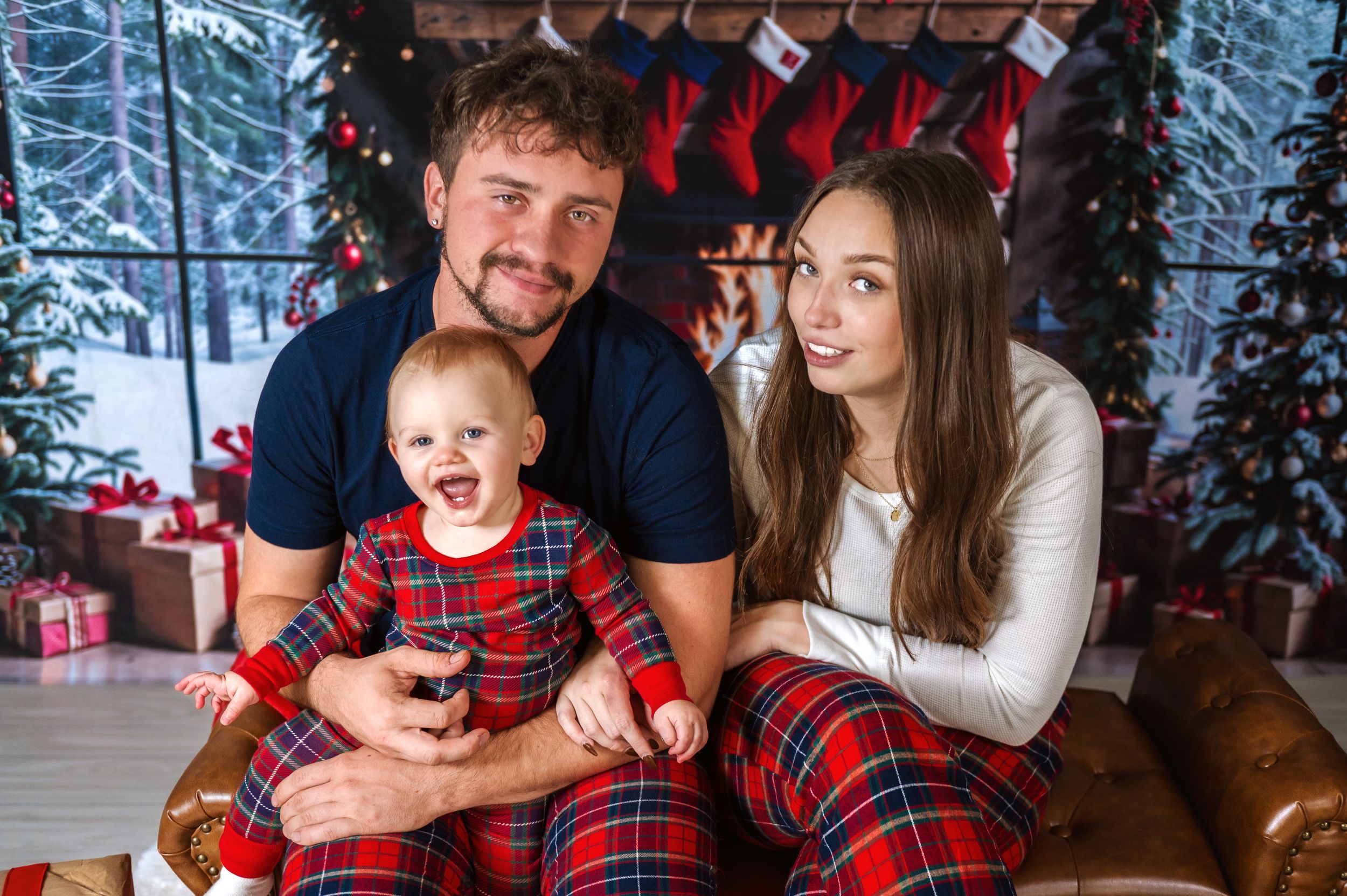 Family wearing matching plaid pajamas poses for Christmas photo with decorated trees and stockings.