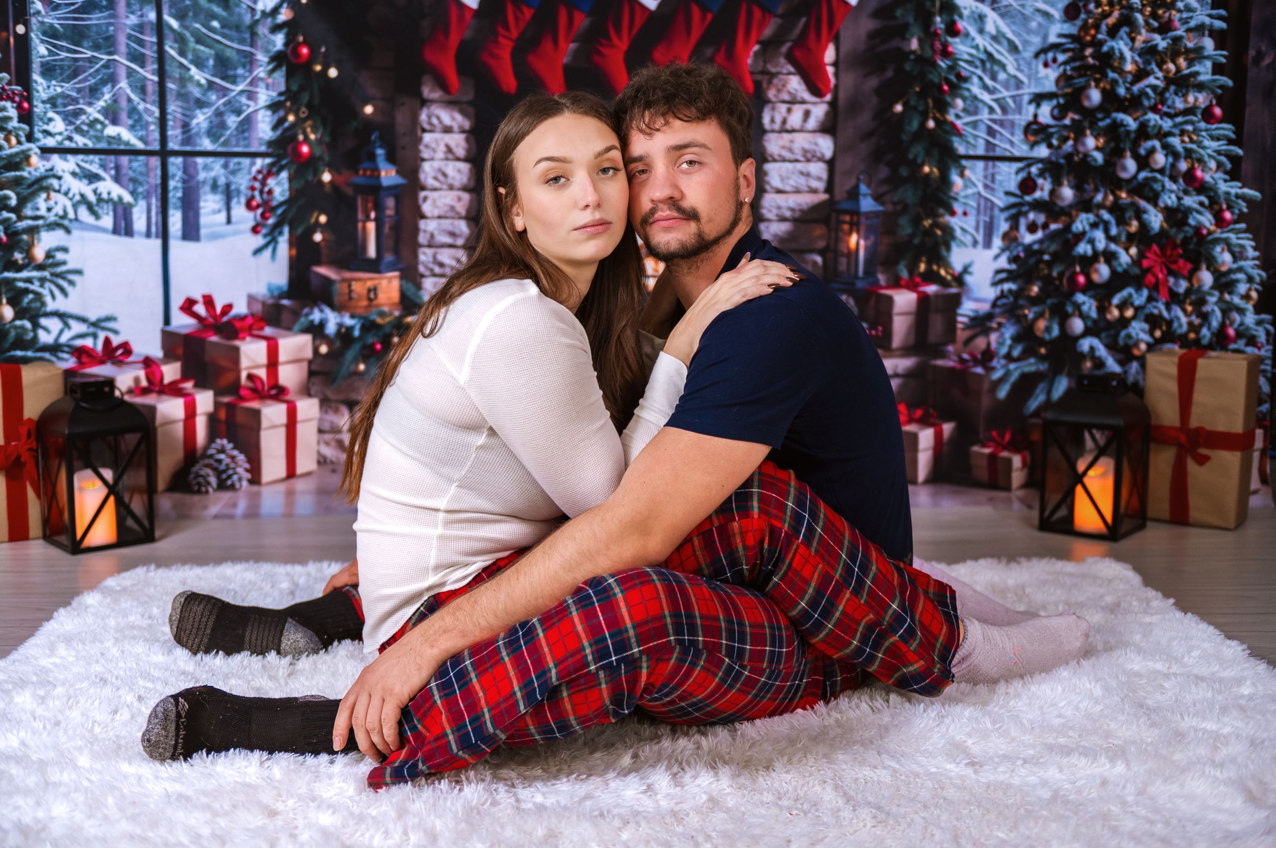 Couple in matching holiday pajamas poses on white fur rug with festive Christmas backdrop.