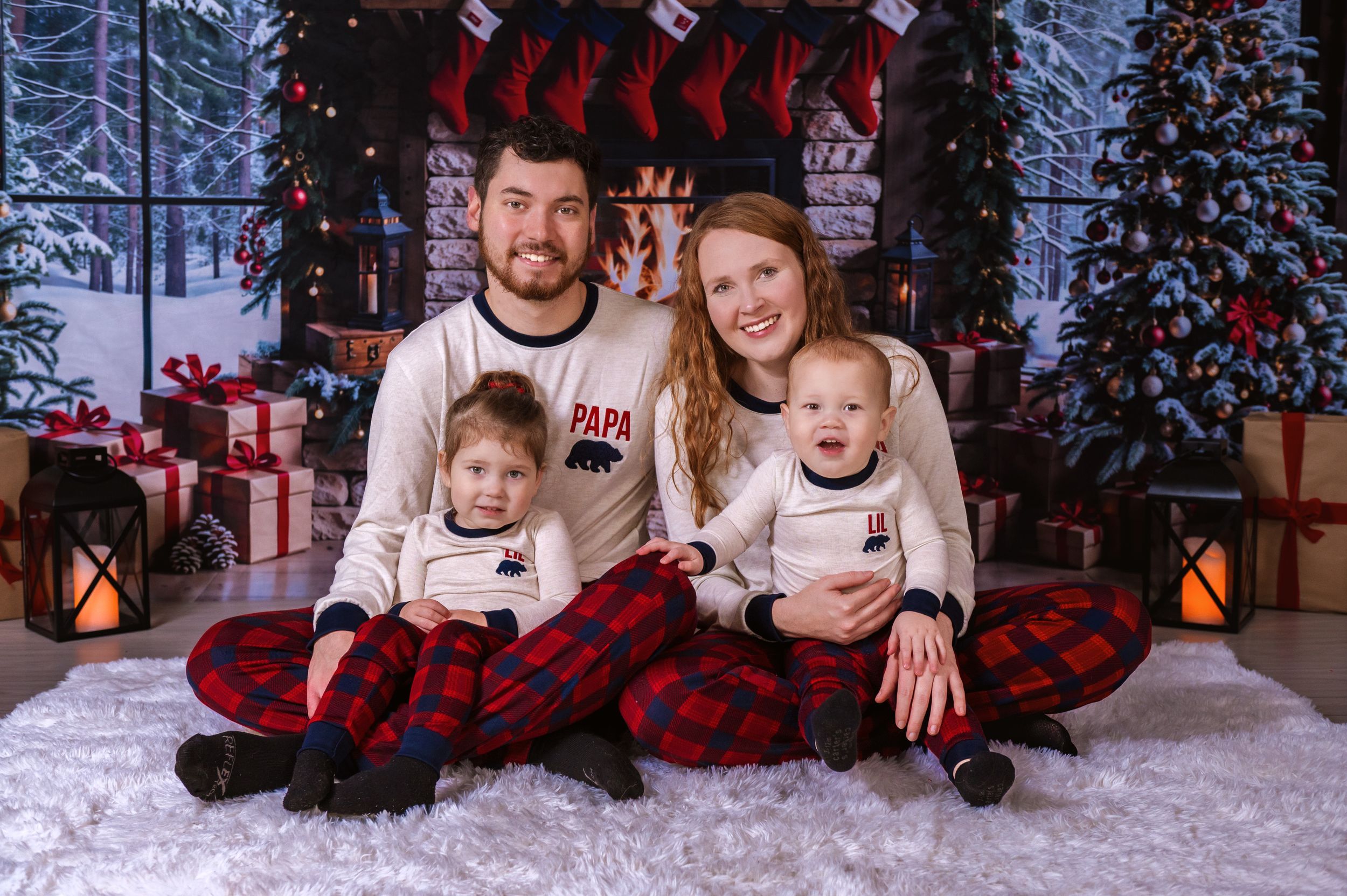 Holiday photoshoot in a cozy Christmas setting with red plaid outfits and snowy backdrop.