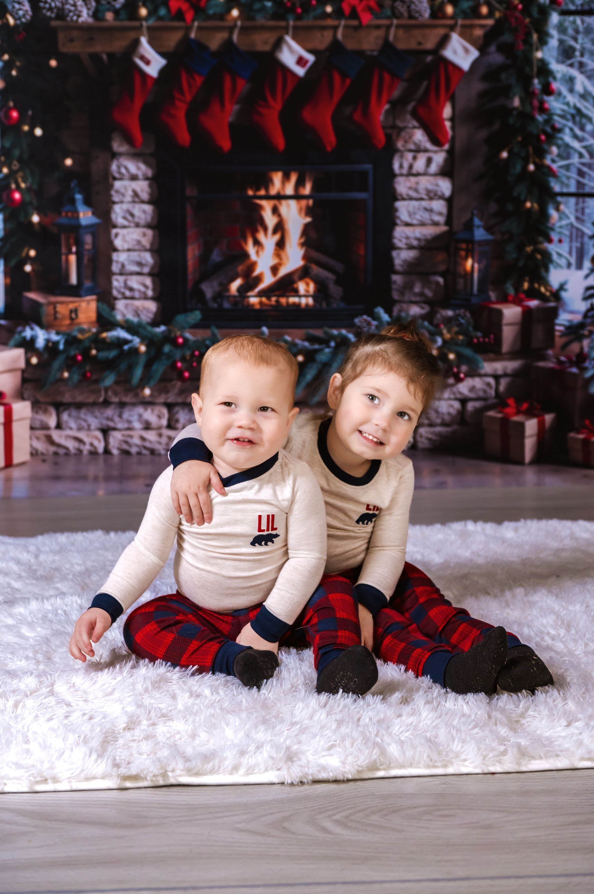 Children lying on white fluffy carpet in front of Christmas fireplace setting.