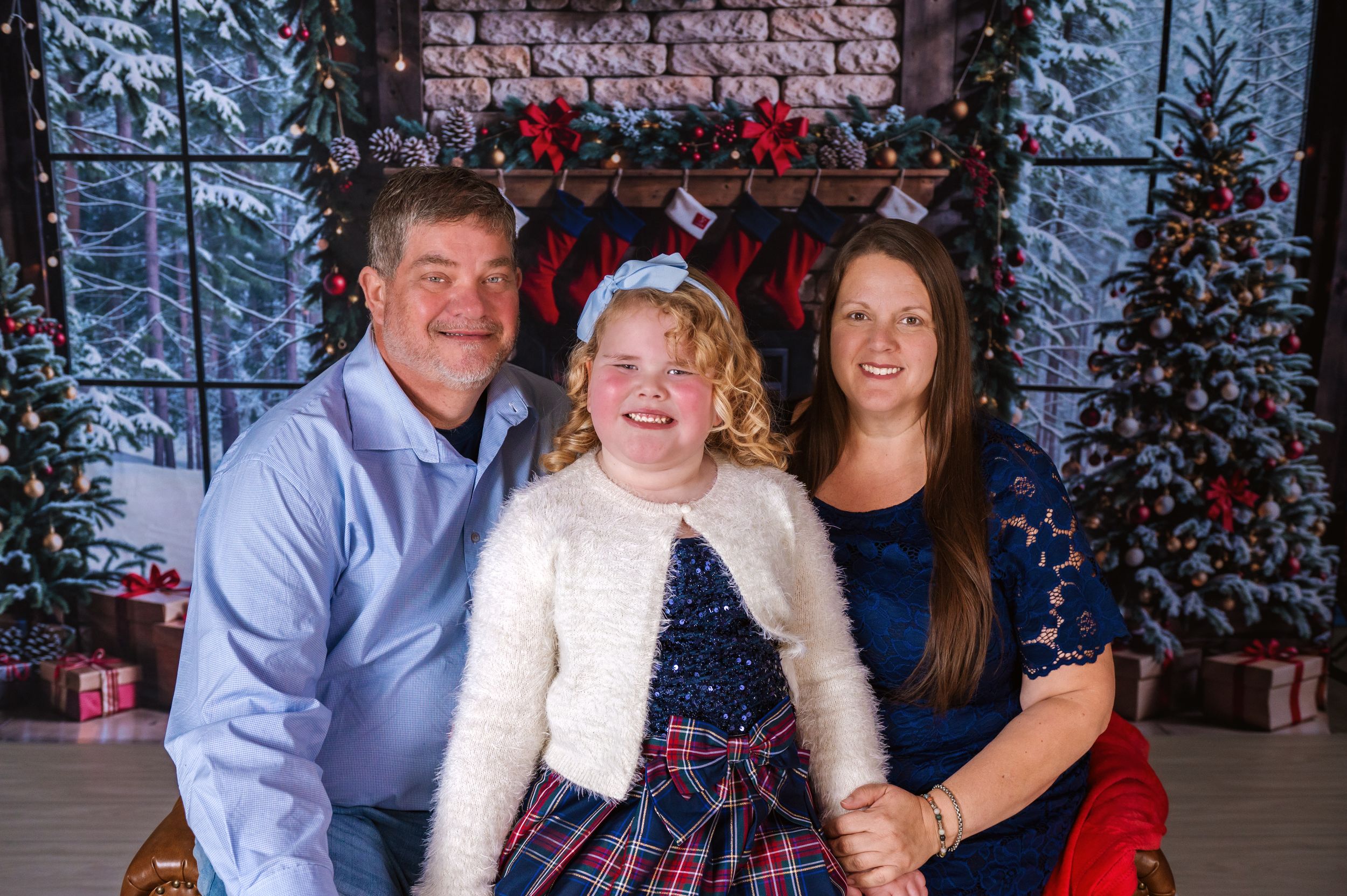 Three people pose for holiday portrait with festive backdrop of red stockings and Christmas trees.