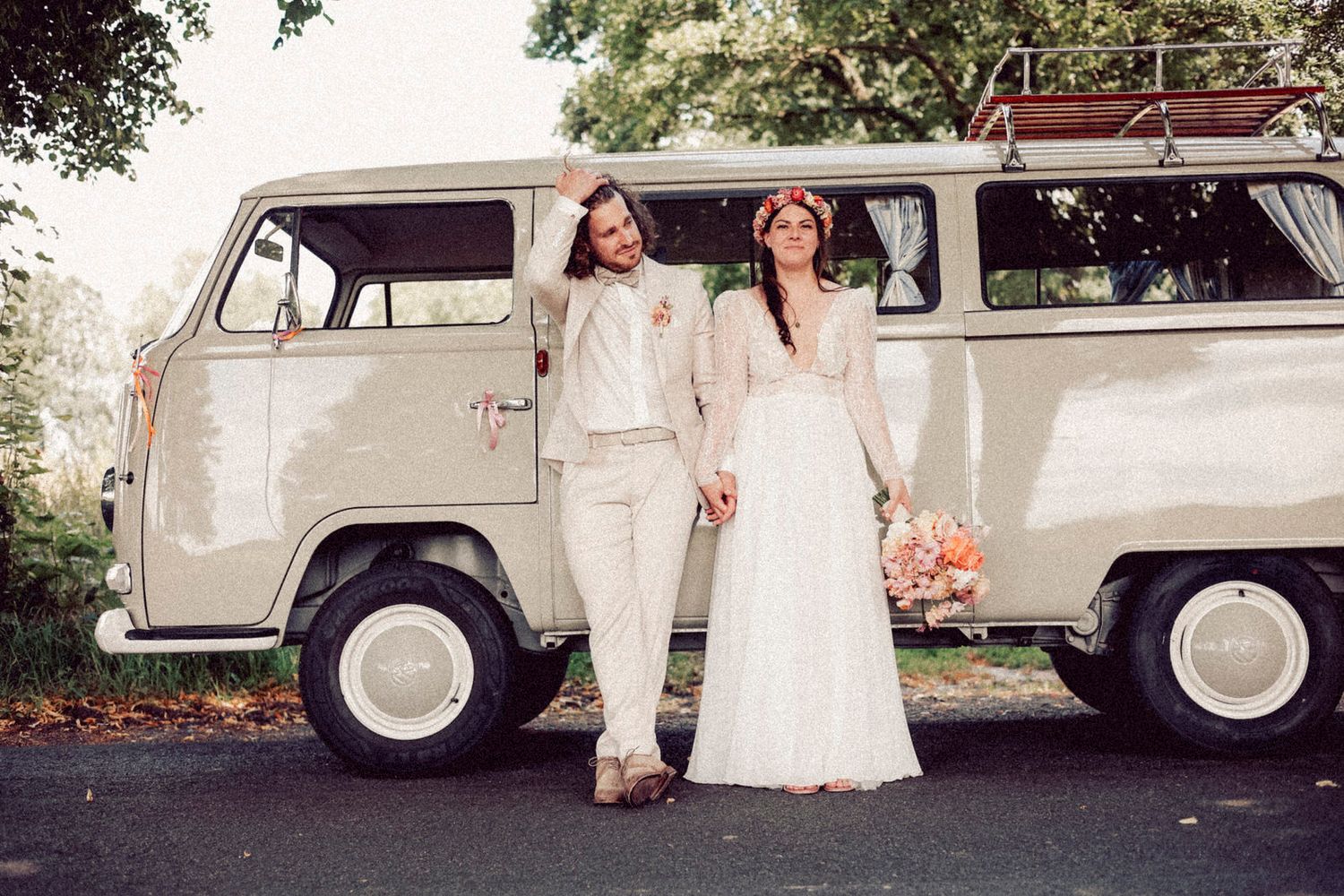 A classic cream VW bus serves as a wedding backdrop with a couple in white posing beside it holding flowers. Bulli Käthe.