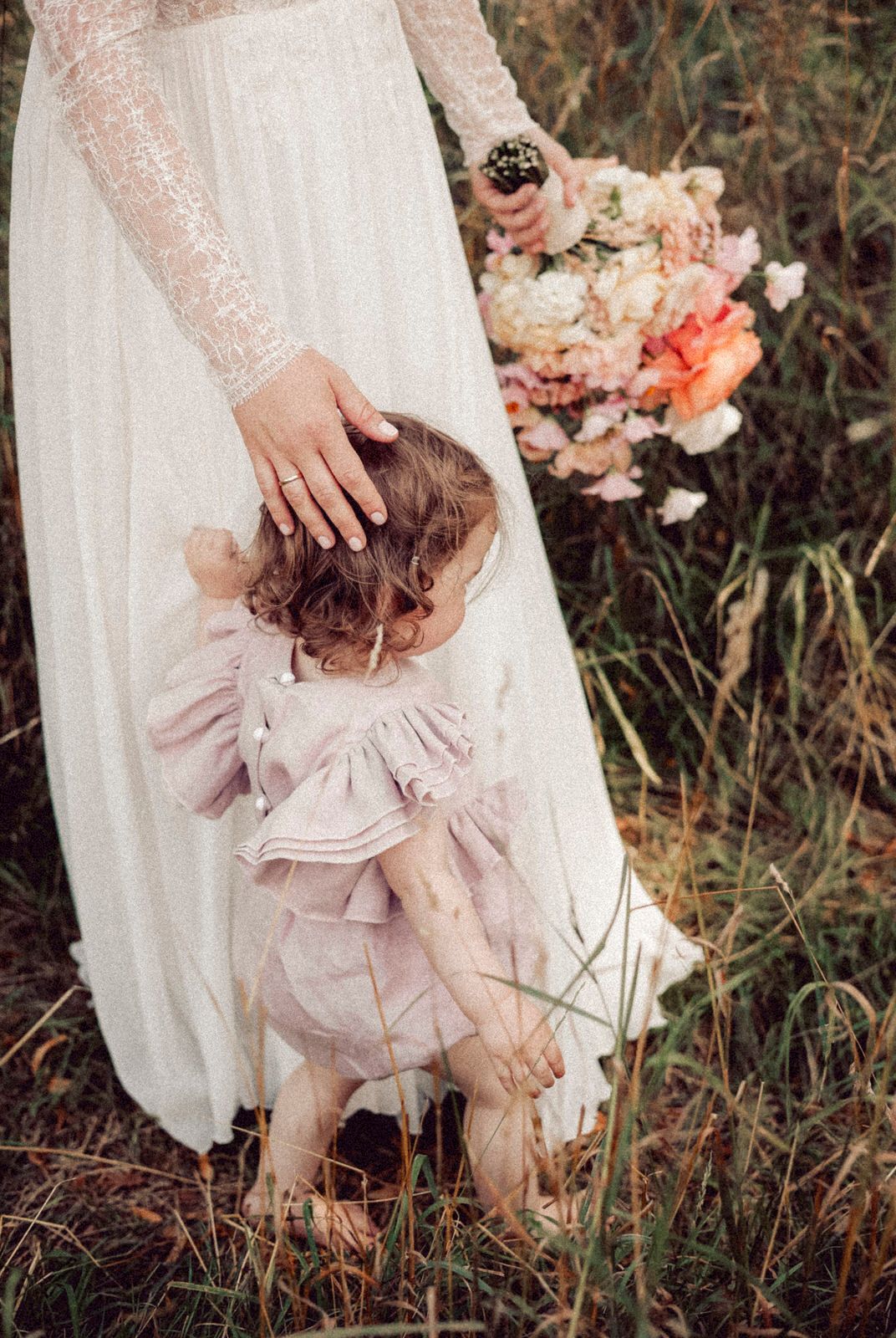 A bride in a white lace wedding dress holds a pink bouquet while standing with a small child in a tulle dress.