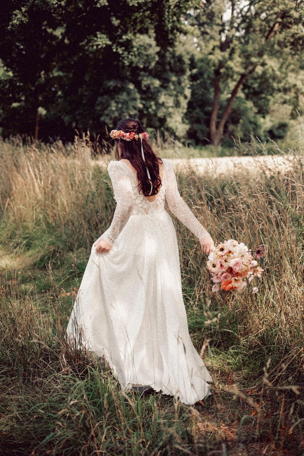 A bride in a bohemian lace wedding dress holds a pink bouquet while walking through tall grass in a natural setting.