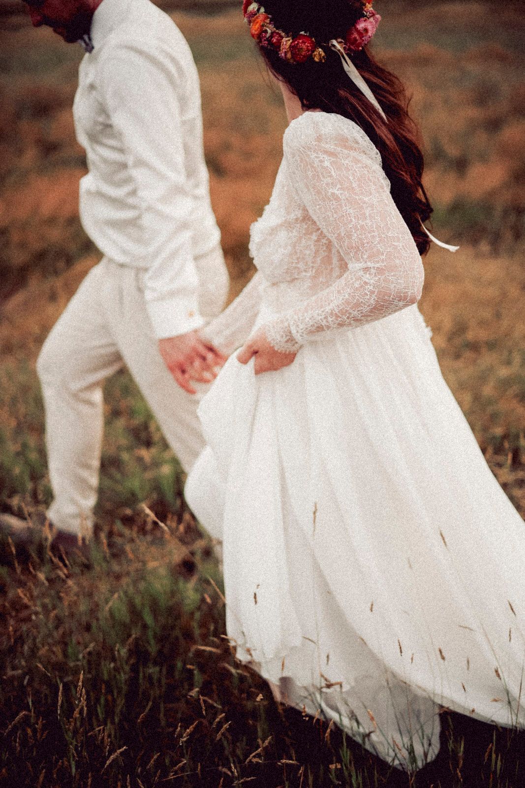 A couple in white wedding attire walks hand in hand through a rustic field, with the bride wearing a flower crown.
