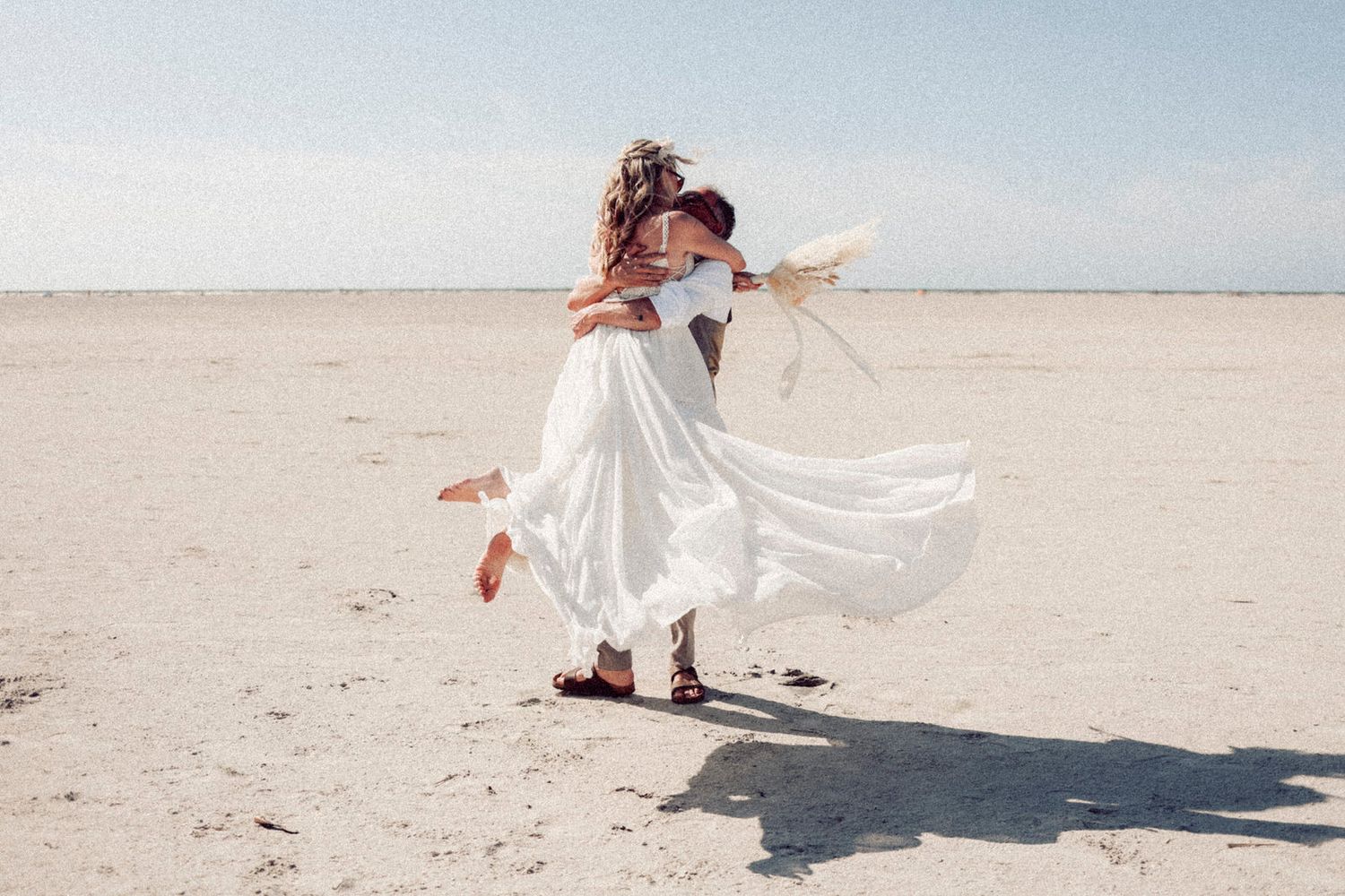 Figure in flowing white dress twirls gracefully on empty beach under bright sunlight with wind catching the fabric. St. Peter Ording.