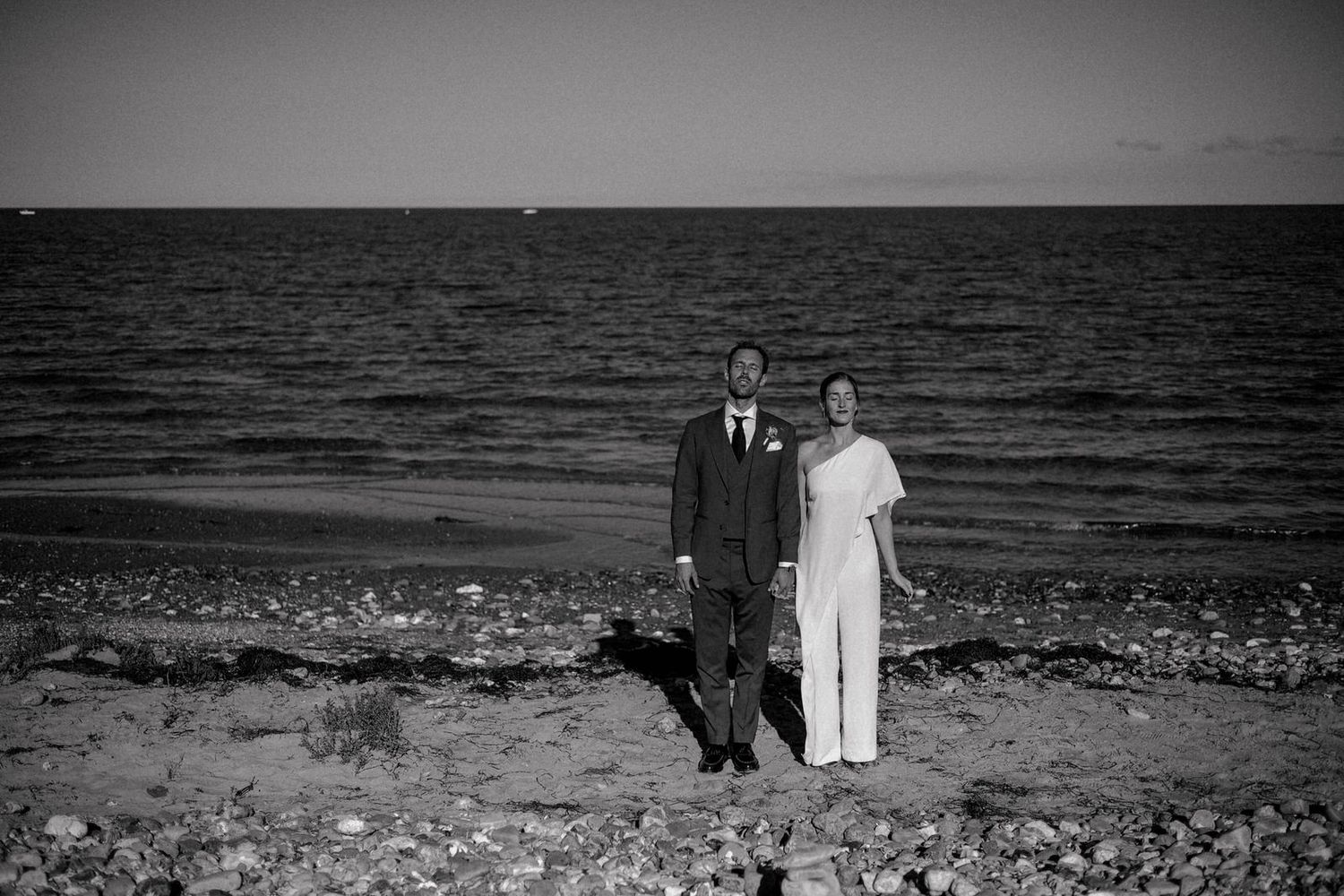 A couple in formal attire stands on a rocky beach shore with the ocean and horizon visible in black and white.