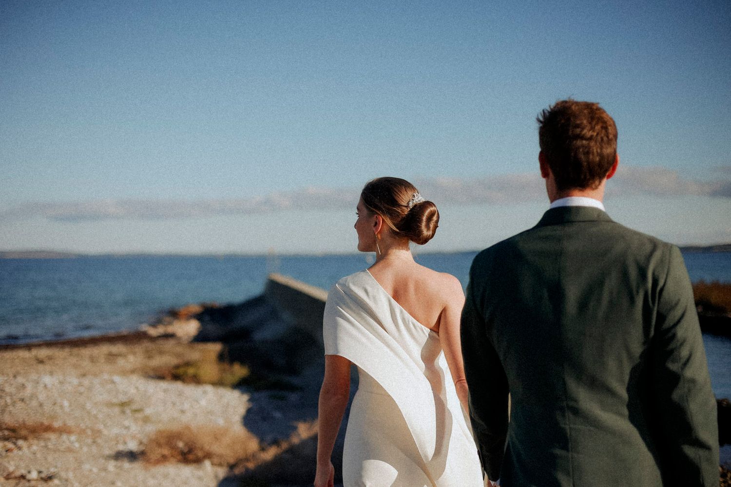 A couple in wedding attire stands together on a rocky coastal cliff overlooking the ocean on a clear sunny day.
