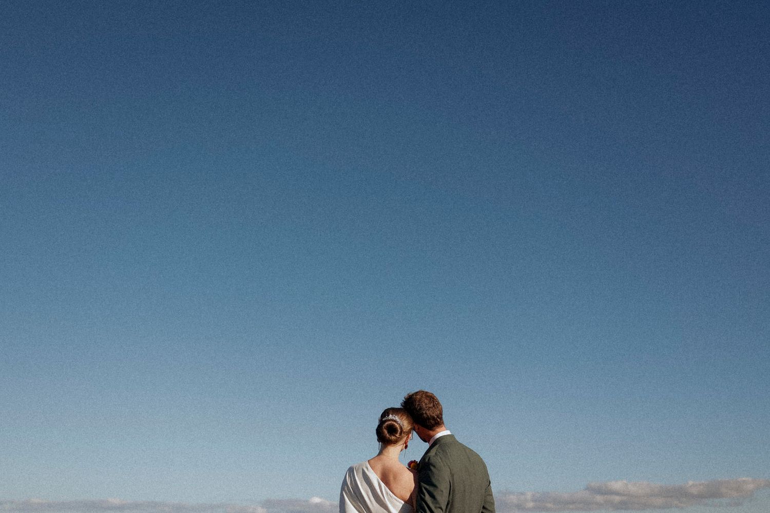 A couple stands close together against a clear blue sky on their wedding day.