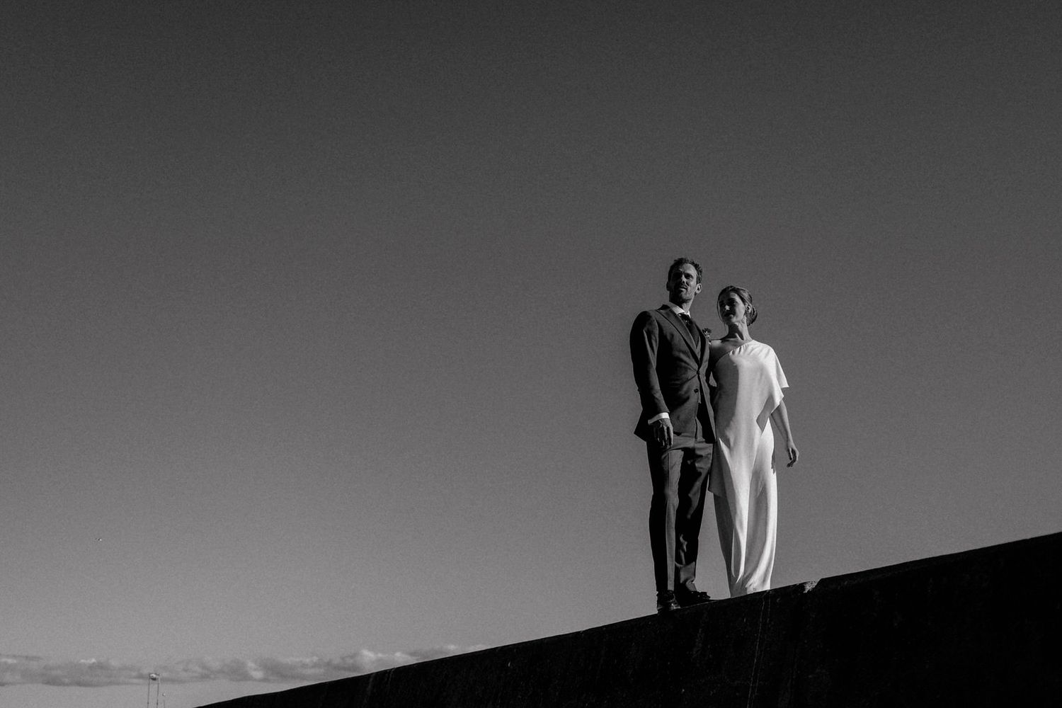 Artistic black and white photo of a couple standing atop a hill silhouetted against a dramatic sky.