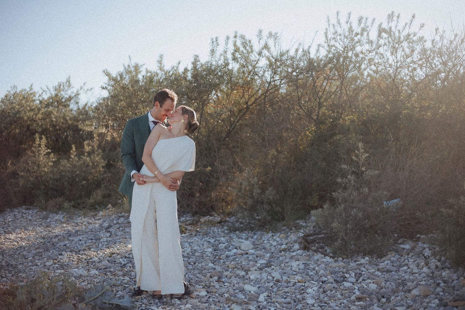 A couple embraces on a rocky path surrounded by desert brush during golden hour sunset.