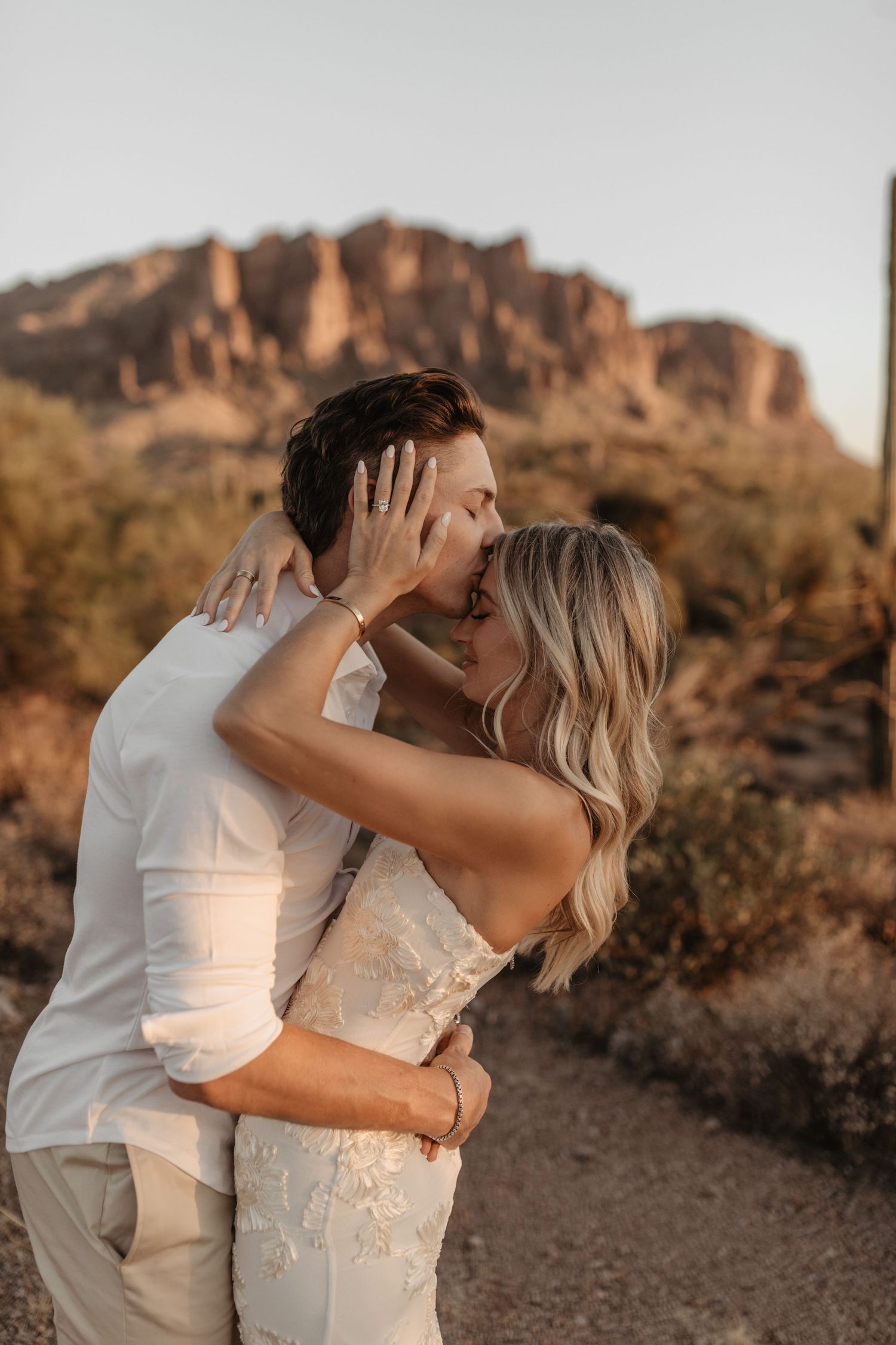 Romantic desert engagement photoshoot featuring a couple embracing at sunset with red rock formations in the background.