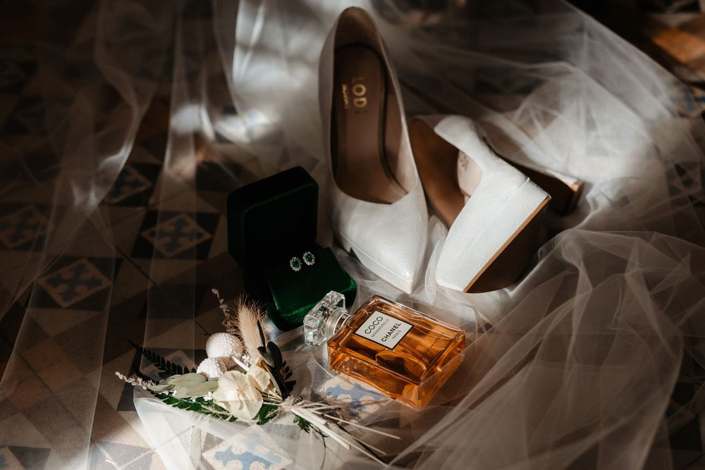 Elegant bridal flat lay featuring white satin heels, Chanel perfume, green velvet ring box, and floral bouquet on lace veil.