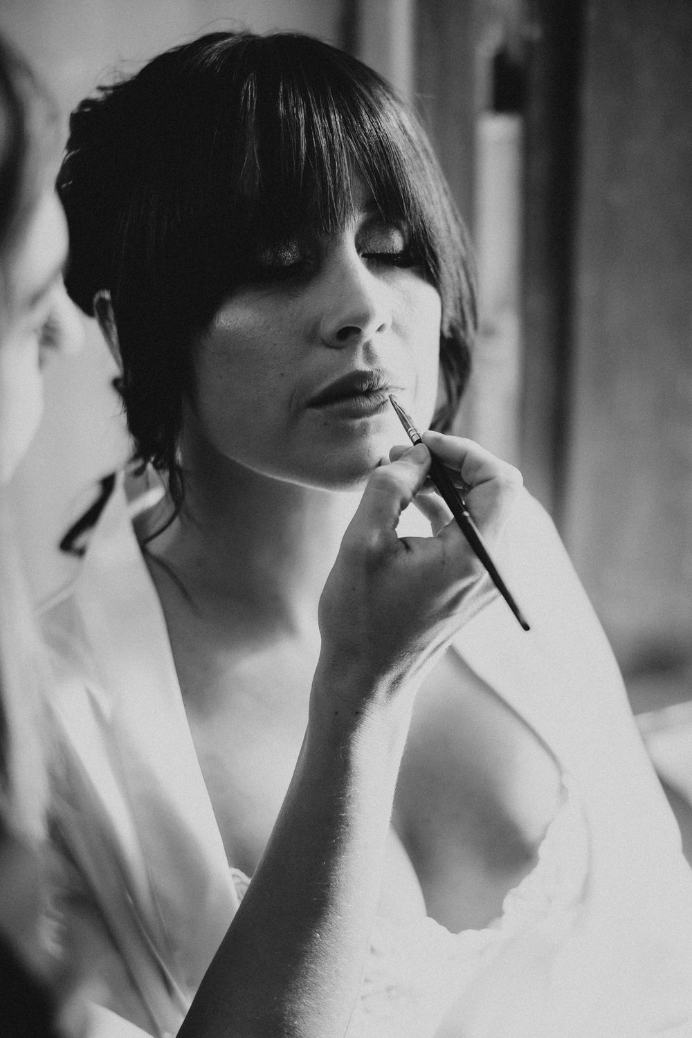 Black and white photo of a bride having lipstick applied by a makeup artist during wedding preparations.