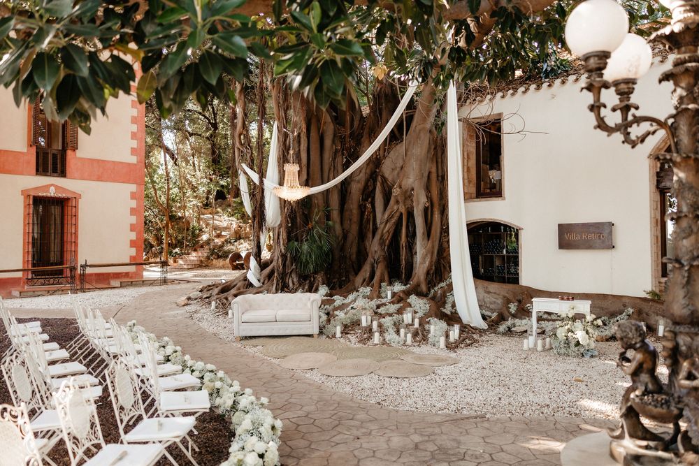 Elegant outdoor wedding ceremony setup with white chairs and floral decor beneath a large banyan tree at a villa.