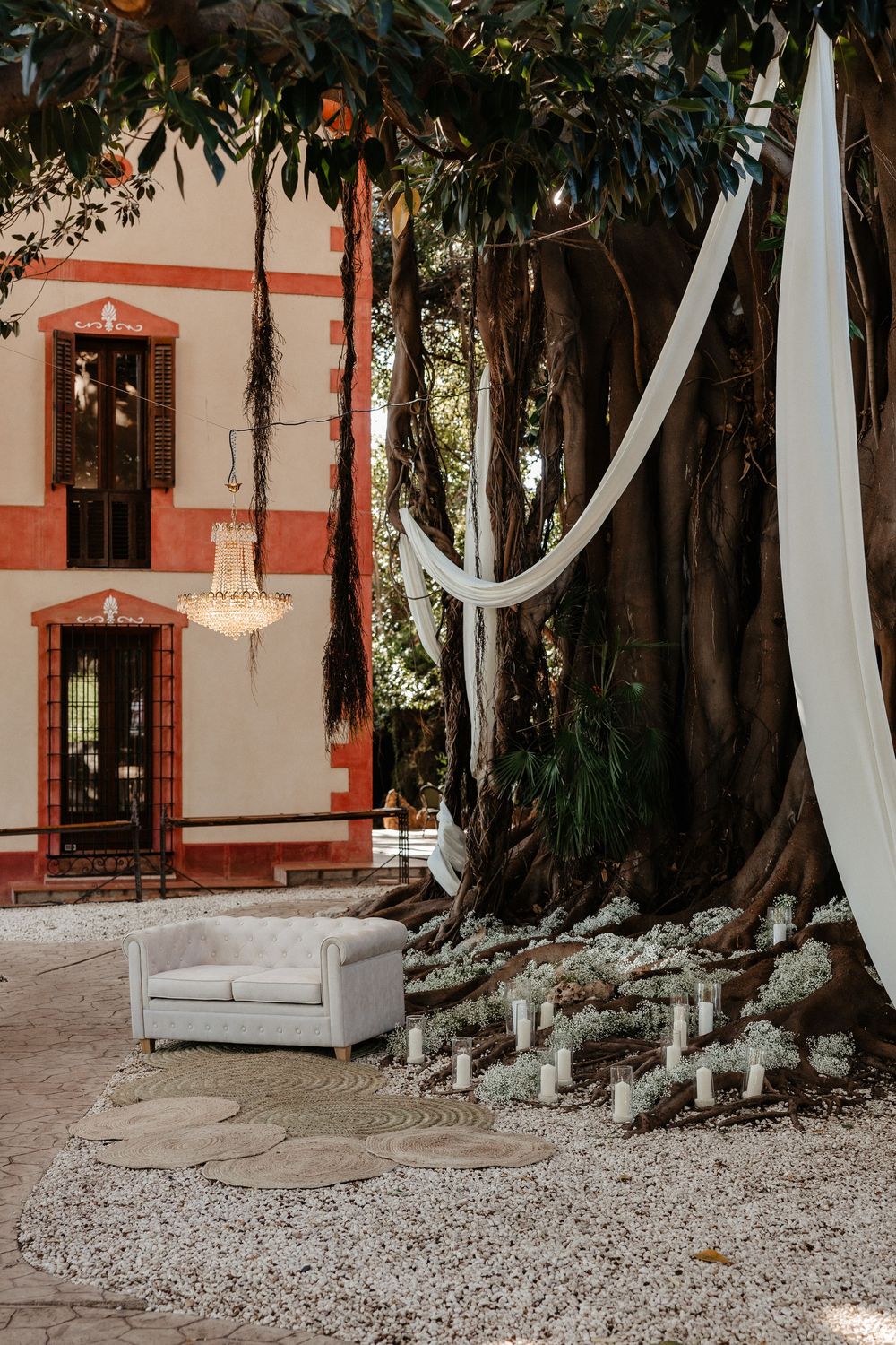 Elegant outdoor wedding setup with white sofa, crystal chandelier, candles, and white draping around a large tree.
