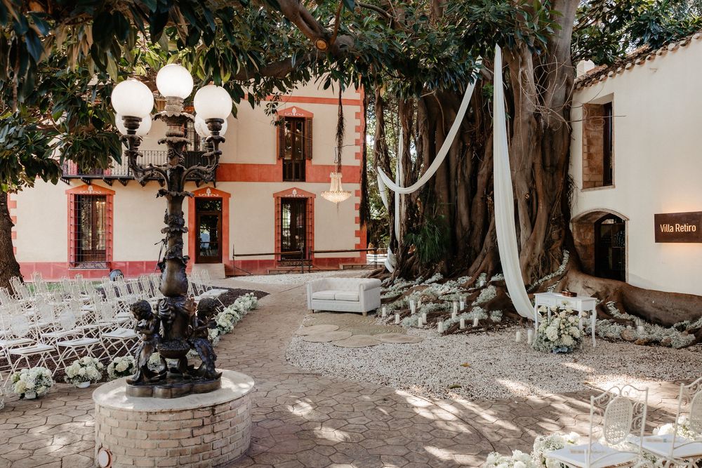 Elegant outdoor wedding venue with white chairs, ornate lamp post, and large tree draped in ribbons beside a historic villa.