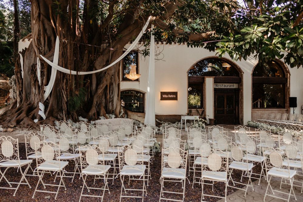 Elegant outdoor wedding ceremony setup with white chairs arranged under a large tree near a white building entrance.