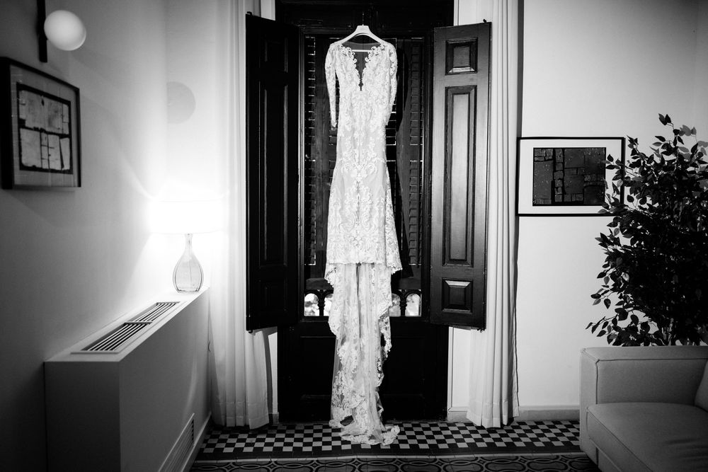Black and white photo of an elegant lace wedding dress hanging on a door in a classic interior room.