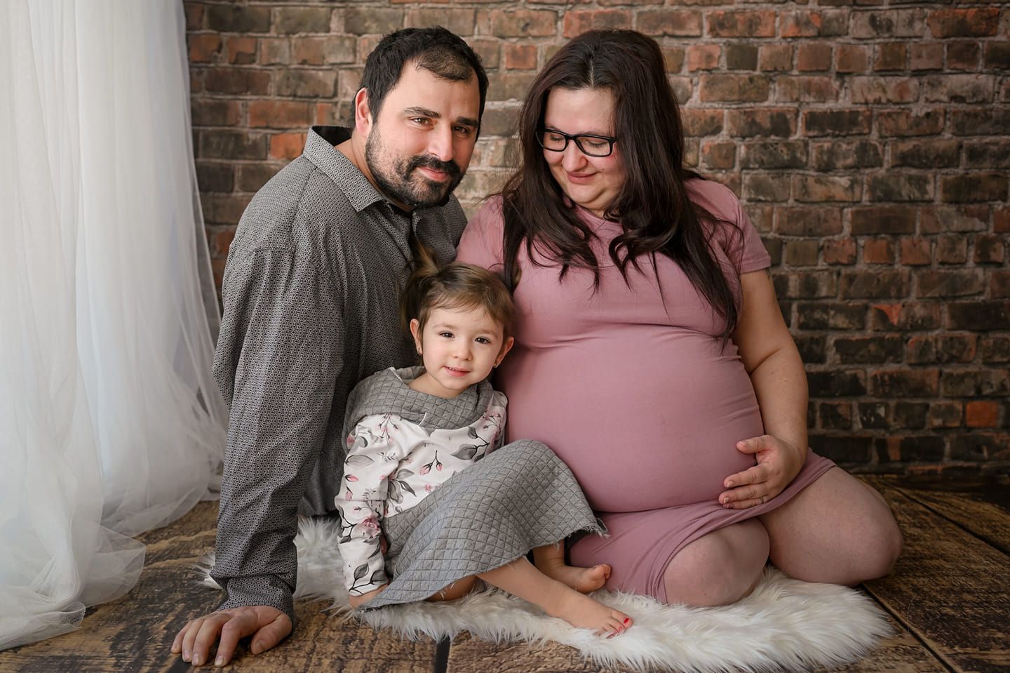 Maternity photo session in a cozy setting with exposed brick wall and soft white draping.