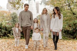 Family in coordinated neutral outfits walking through autumn leaves with city skyline behind them.