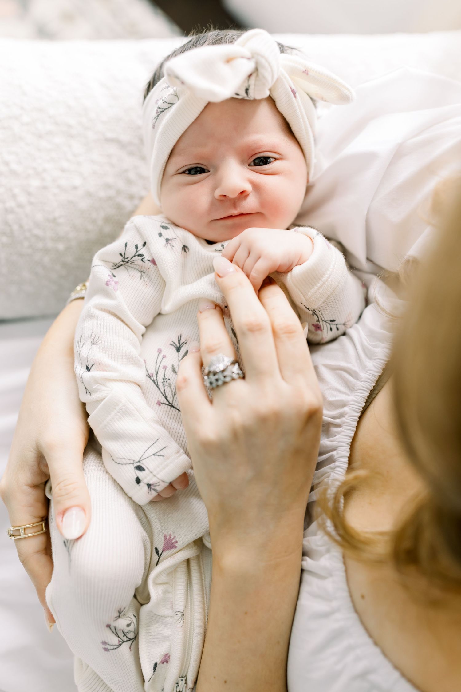 Baby wearing a white bow headband and floral outfit smiles while lying on white bedding.
