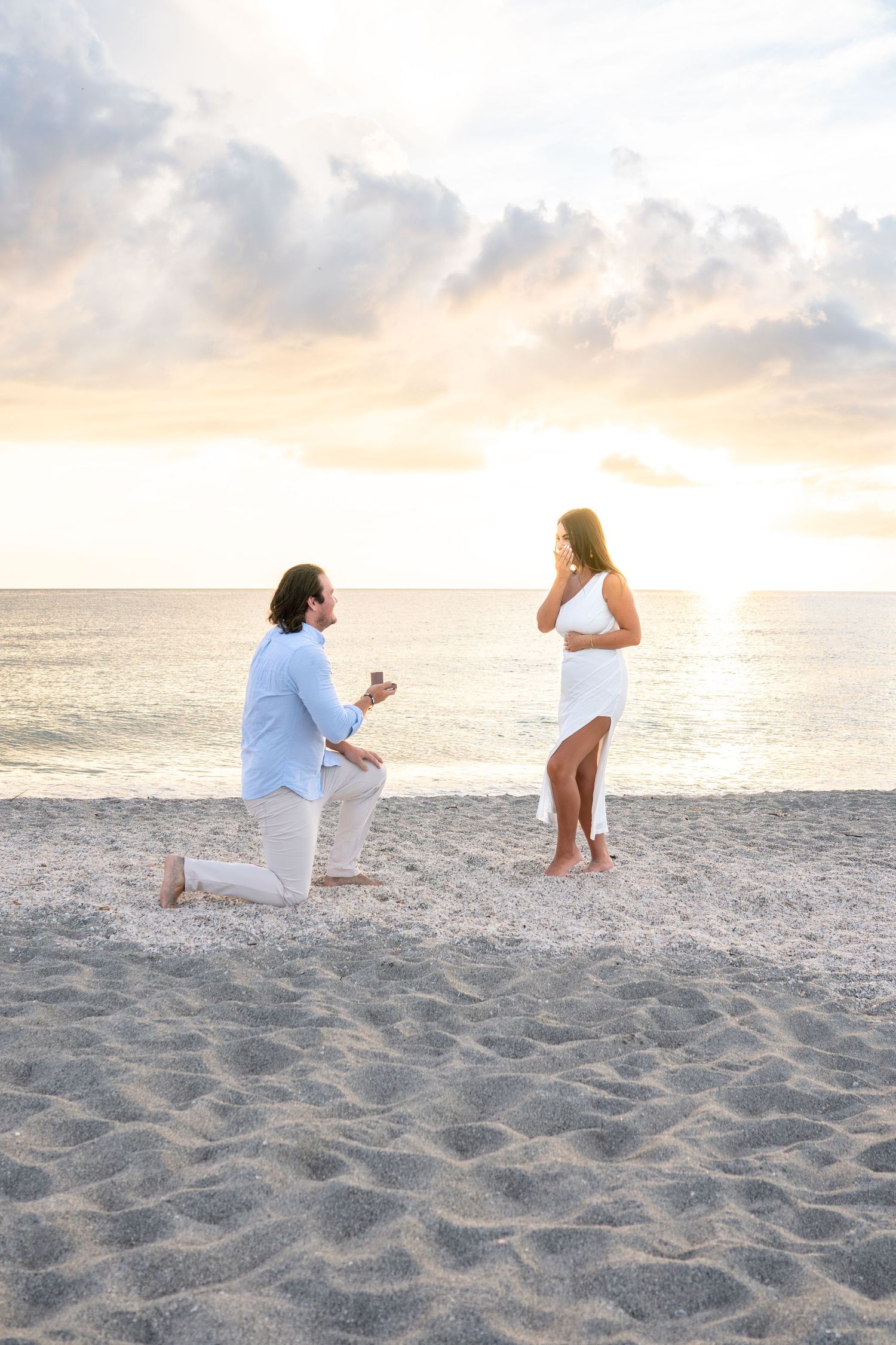 Beach marriage proposal at sunset with man on one knee before woman.