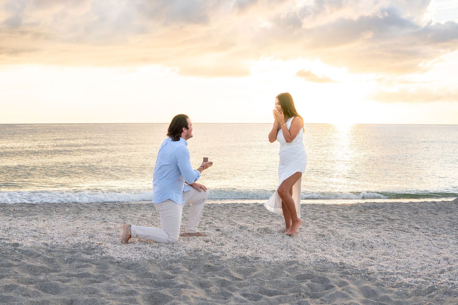 Man proposing on one knee to surprised woman on beach at sunset.