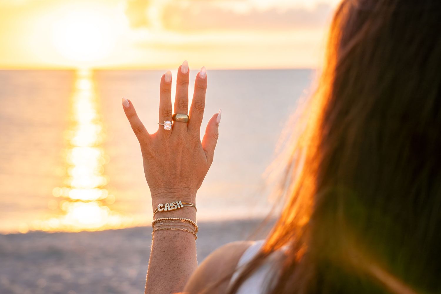 Close-up of womans hand displaying engagement ring and gold bracelet against a stunning ocean sunset.