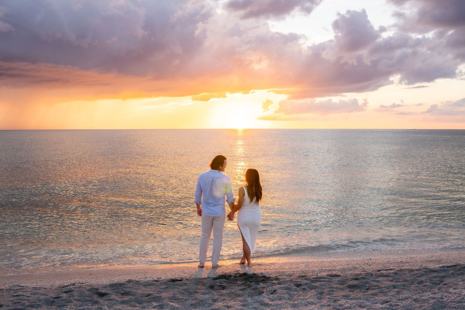 Couple holding hands watching a breathtaking orange sunset over the calm ocean on the beach.